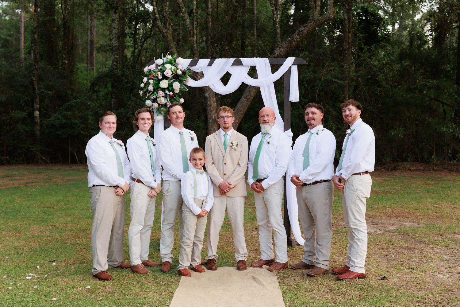 Groomsmen in light khaki suits and white shirts pose with groom under decorative arch in wooded setting.
