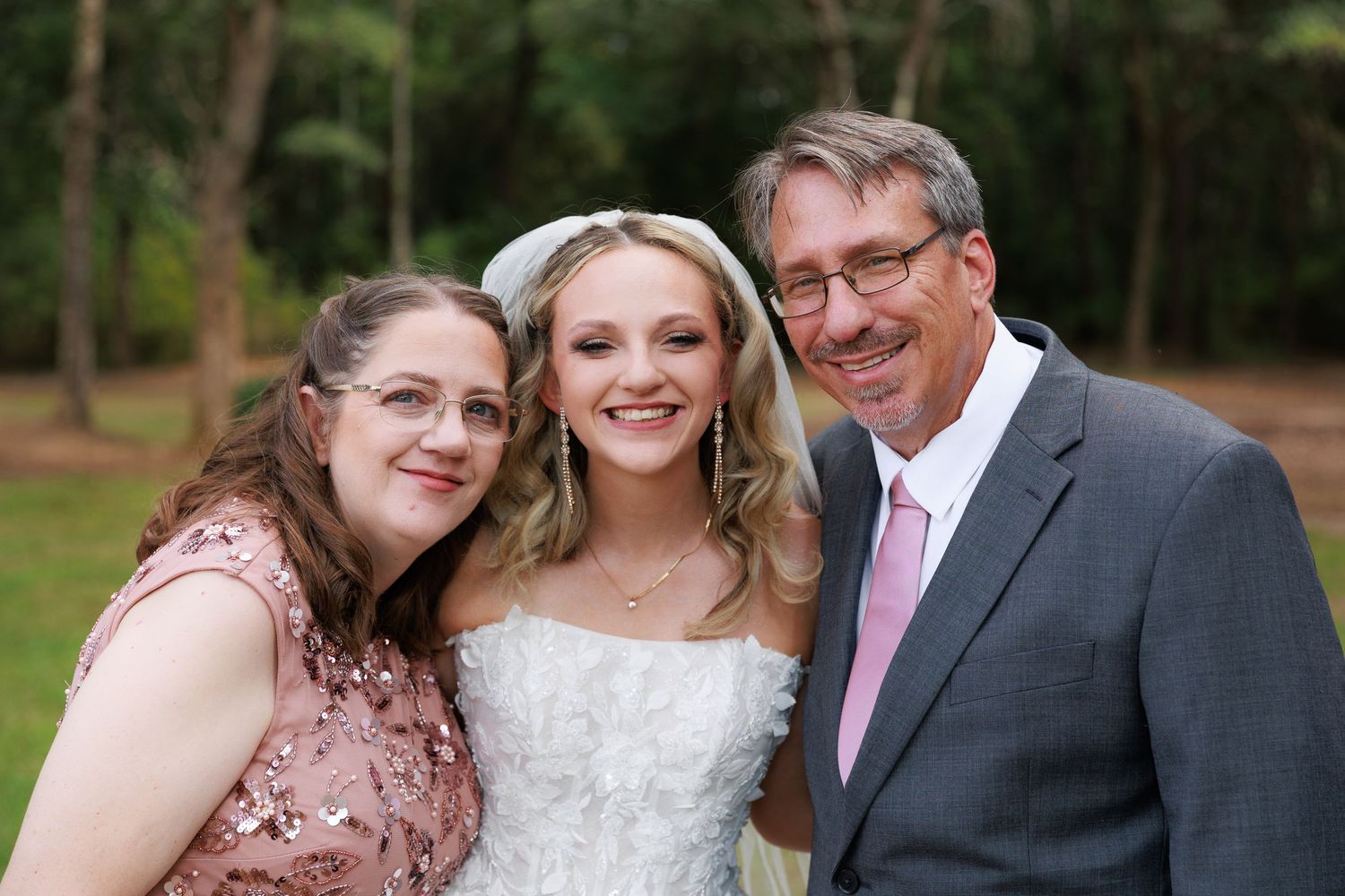 Three people pose together outdoors at a formal event, with a bride in white dress flanked by two guests in pink attire.