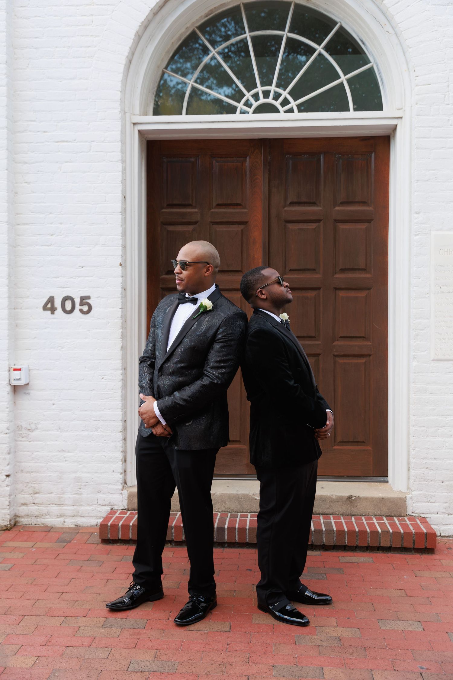Two men in black suits lean against a white brick wall near a door marked 405 on red brick pavement.