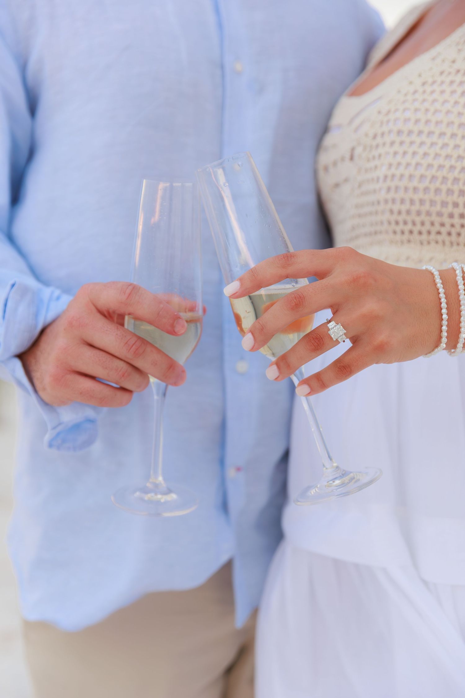 Two people toasting champagne flutes, wearing light blue and white clothing with a diamond ring and bracelet visible.