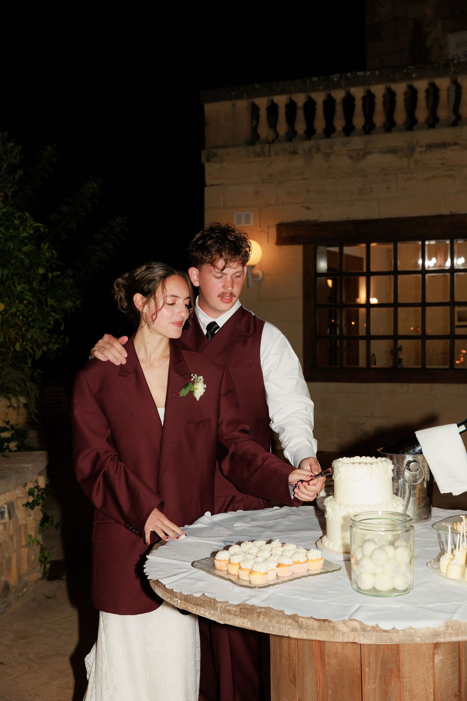 A couple in burgundy attire cuts their wedding cake together at an outdoor evening reception.