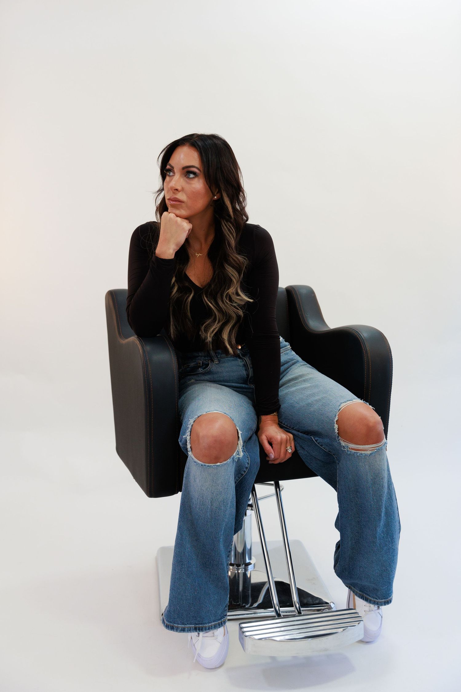 Person in black sweater and distressed denim poses thoughtfully in salon chair.
