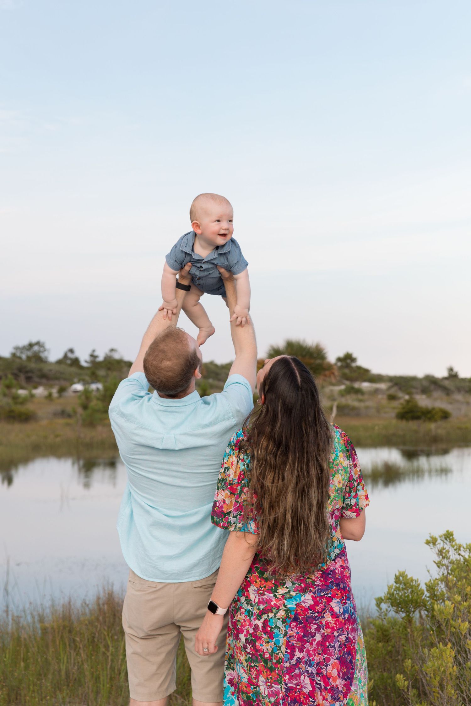 Parents playing with their child against a scenic coastal marsh landscape during golden hour.