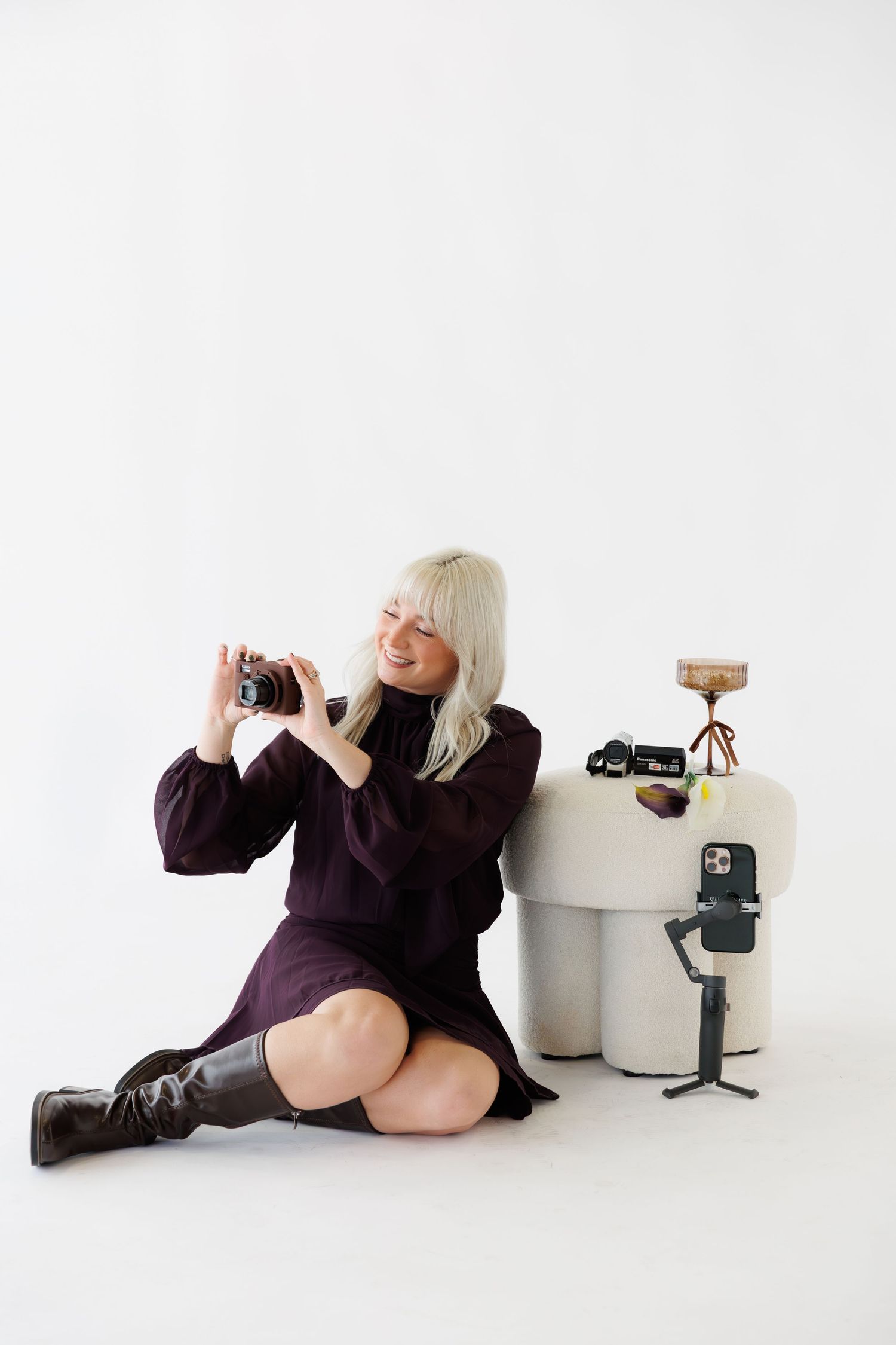 Person in purple dress sitting on floor with camera and cocktail glass in minimalist studio setting.