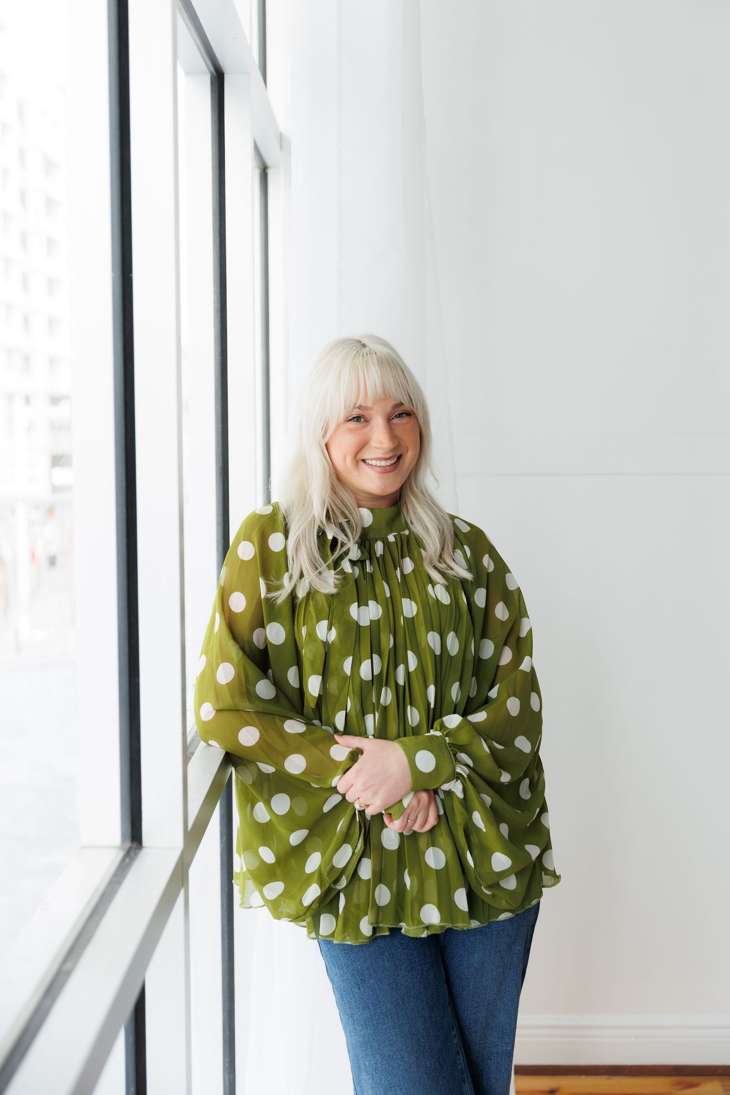 Someone wearing a green polka dot blouse and jeans poses cheerfully by bright windows in a modern space.