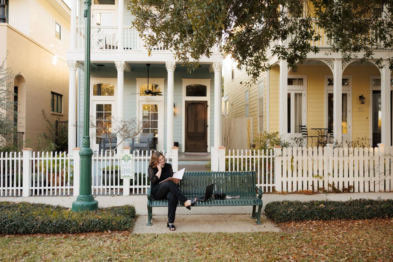 Person sitting on a green bench outside a charming yellow Victorian home with white picket fence.