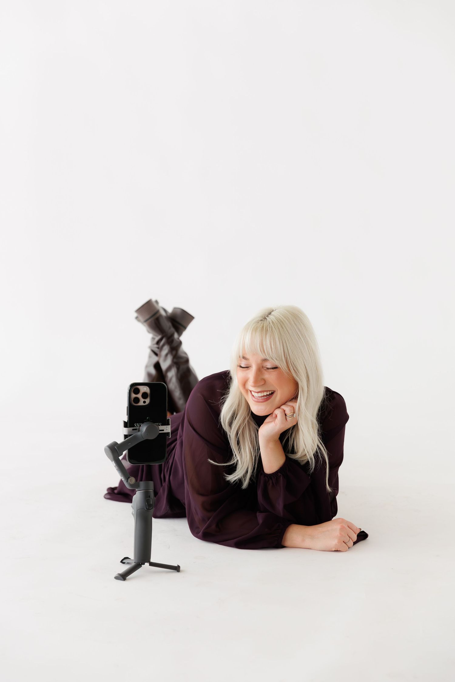 Smiling blonde woman lying on white floor recording herself on a smartphone mounted on a gimbal stabilizer.