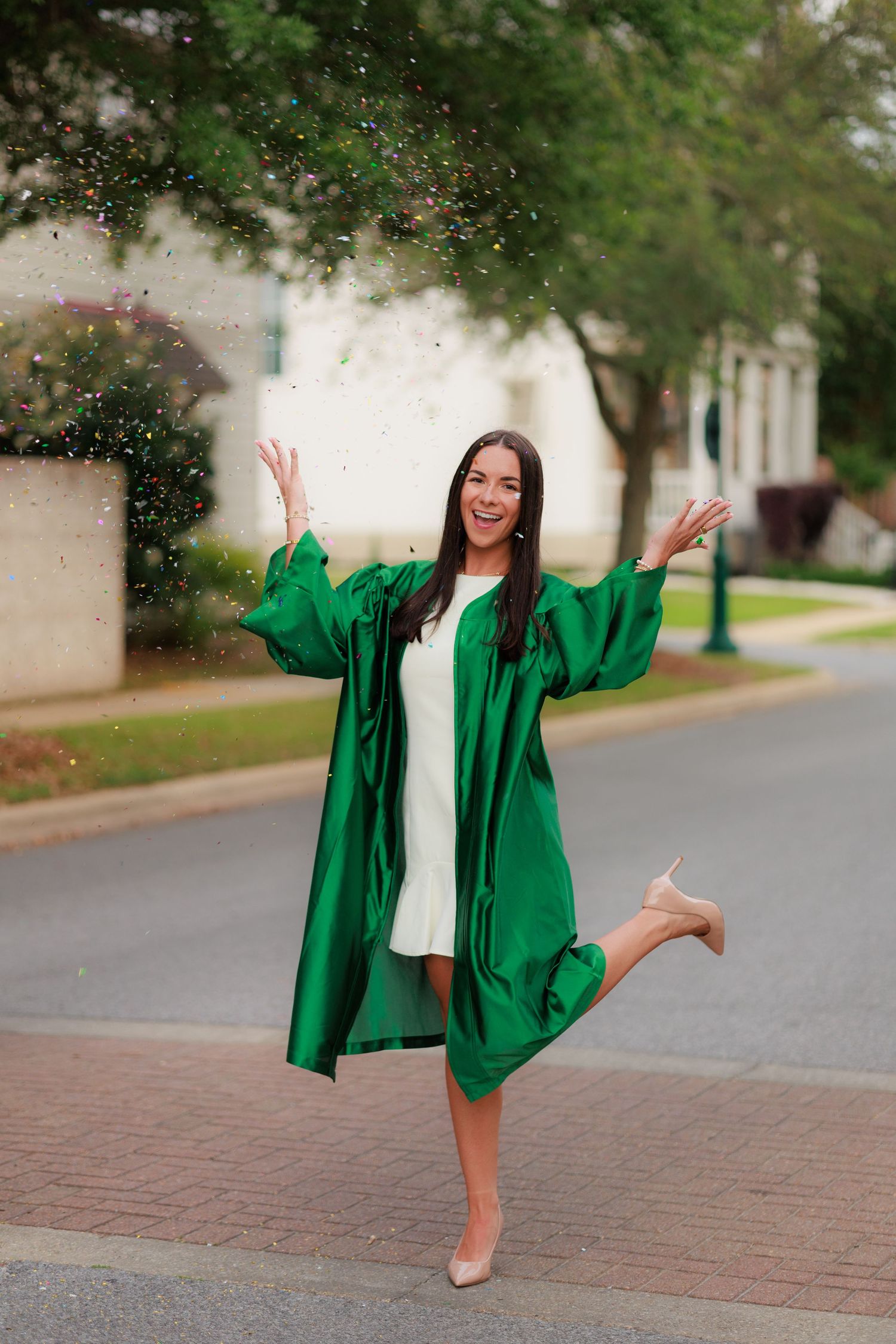 Excited female graduate in green gown celebrating with confetti toss while lifting leg on campus.