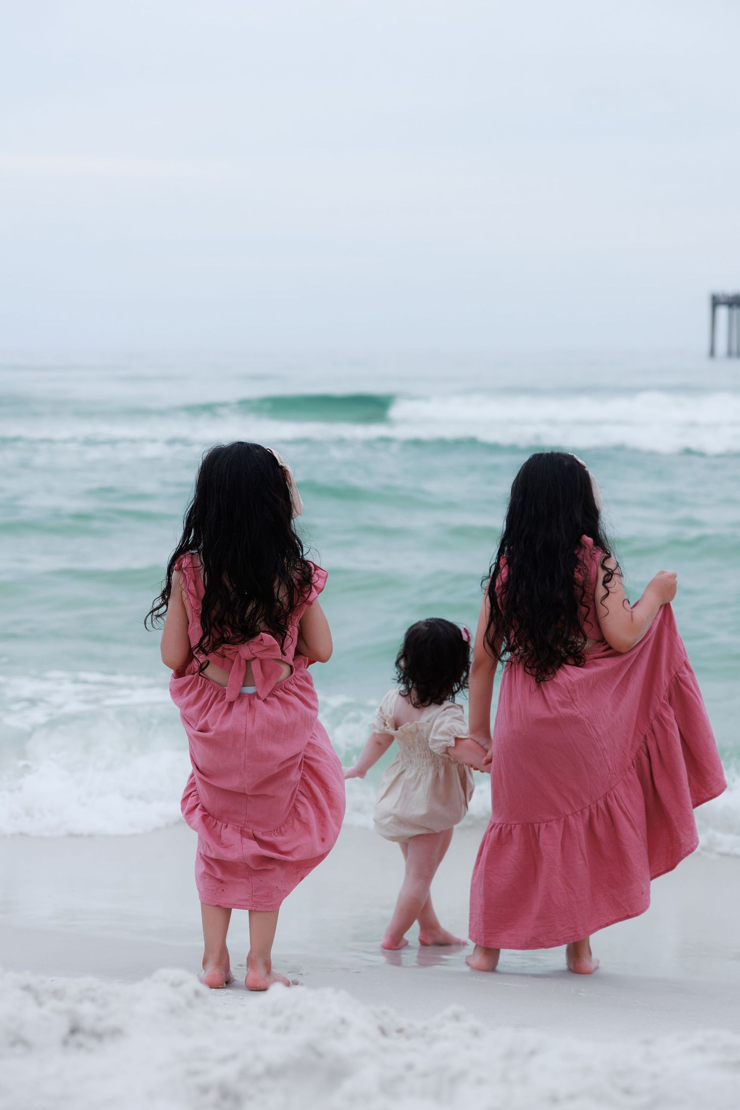 Three girls in pink dresses stand barefoot at the ocean's edge on a beach, viewed from behind.