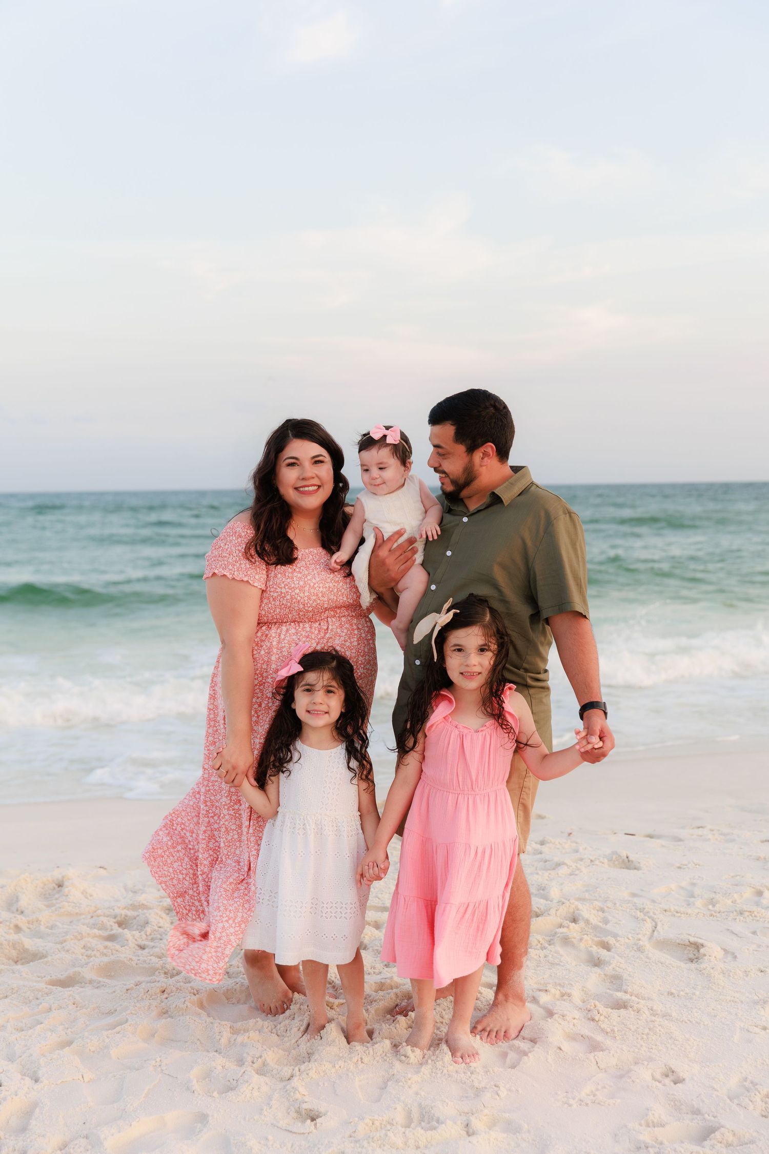 Family of five posing on a sandy beach at sunset, parents with three young daughters in pink and white dresses.
