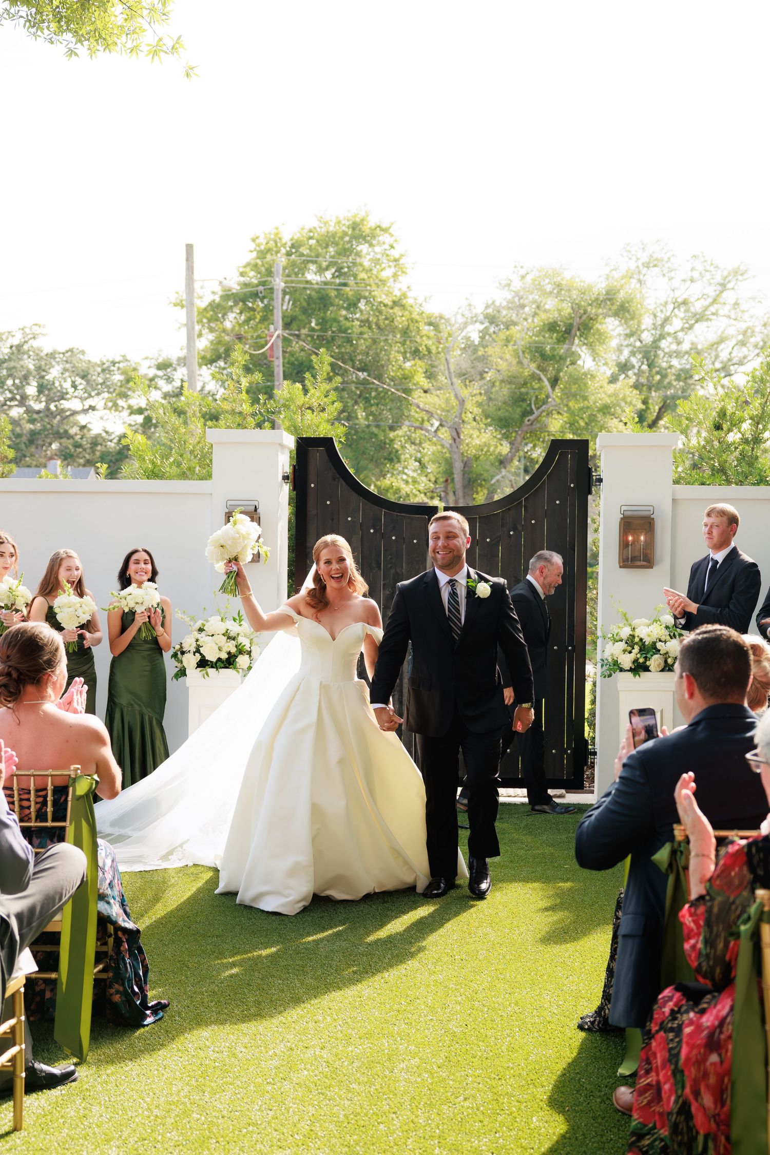 Bride and groom walk back down the aisle after wedding ceremony on lush green lawn with guests applauding.