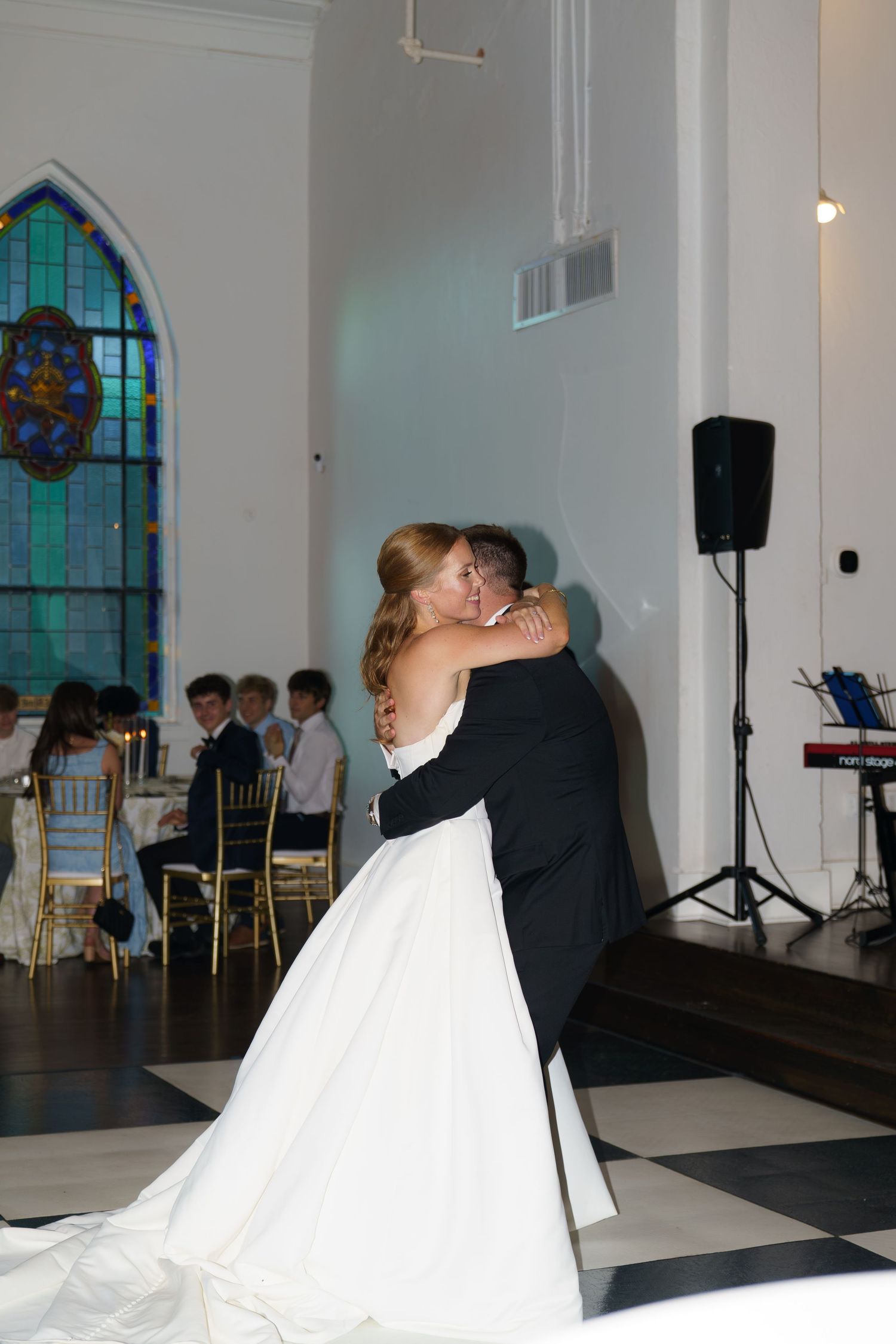 Bride and groom share their first dance in an elegant white church venue with stained glass windows.