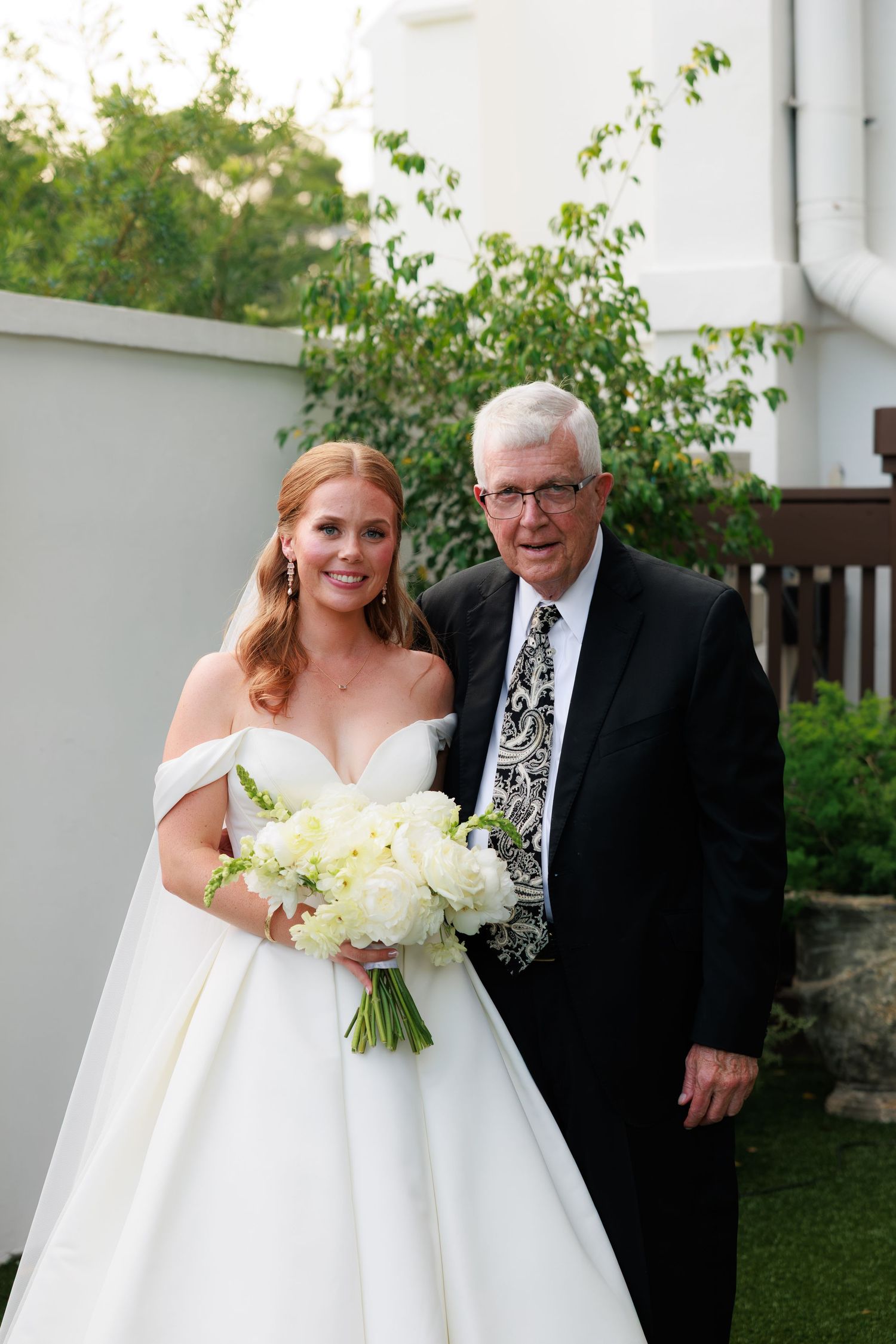 Bride in off-shoulder white gown holding bouquet poses with older gentleman in black suit outdoors.