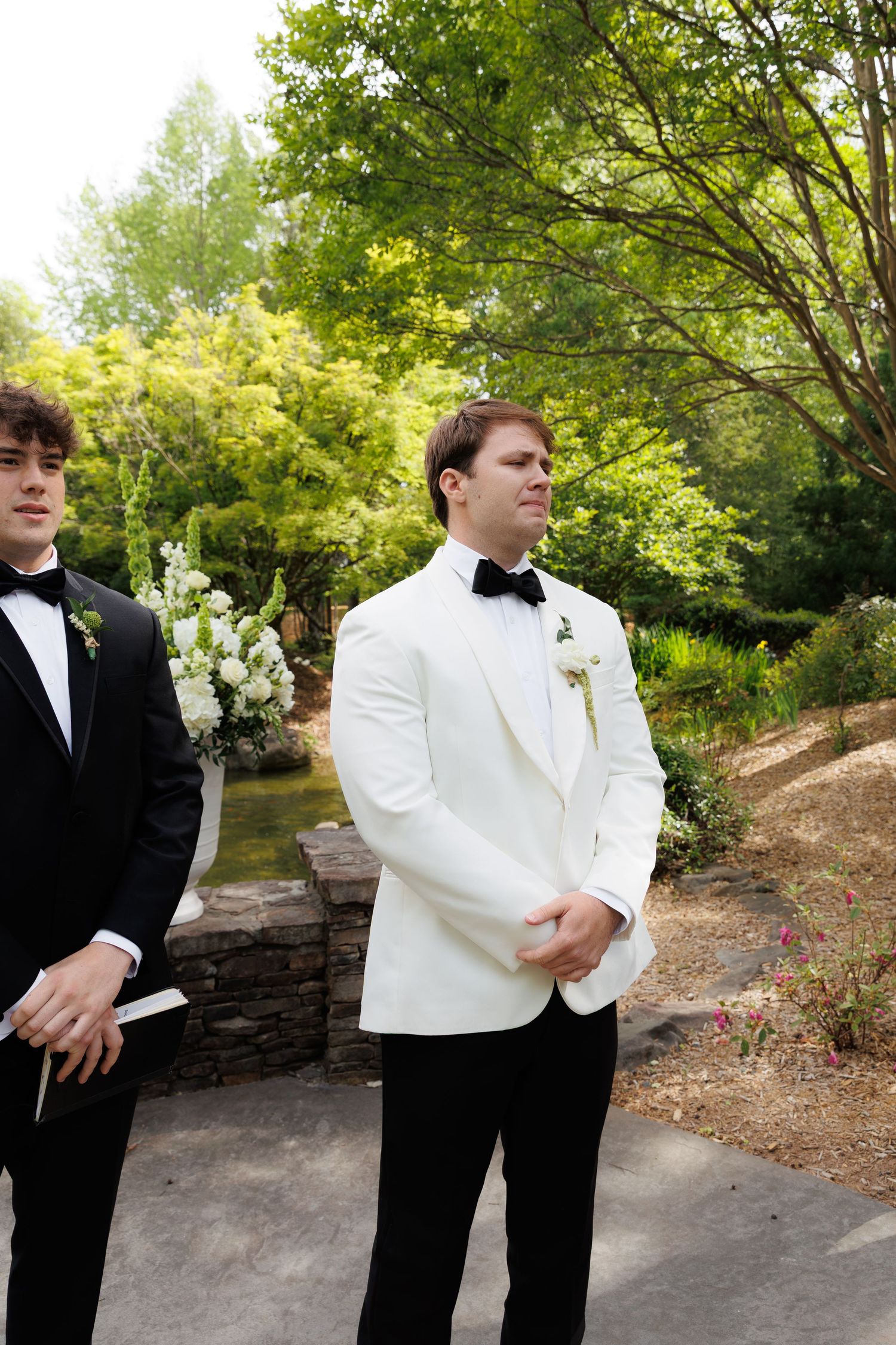 Groom in white tuxedo jacket and black bow tie stands outdoors at wedding ceremony with groomsman beside him.