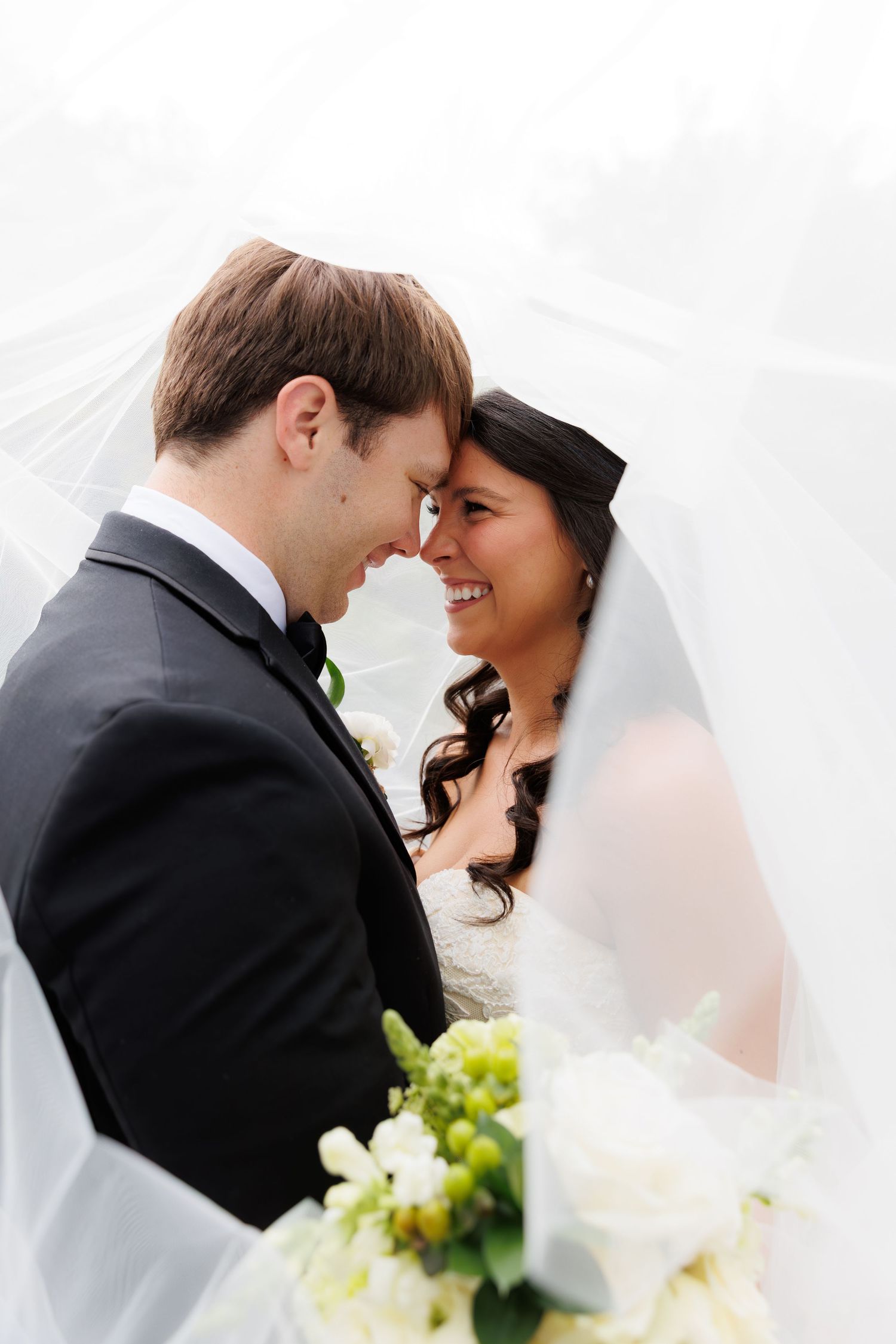 Bride and groom touch foreheads smiling under wedding veil with white floral bouquet.