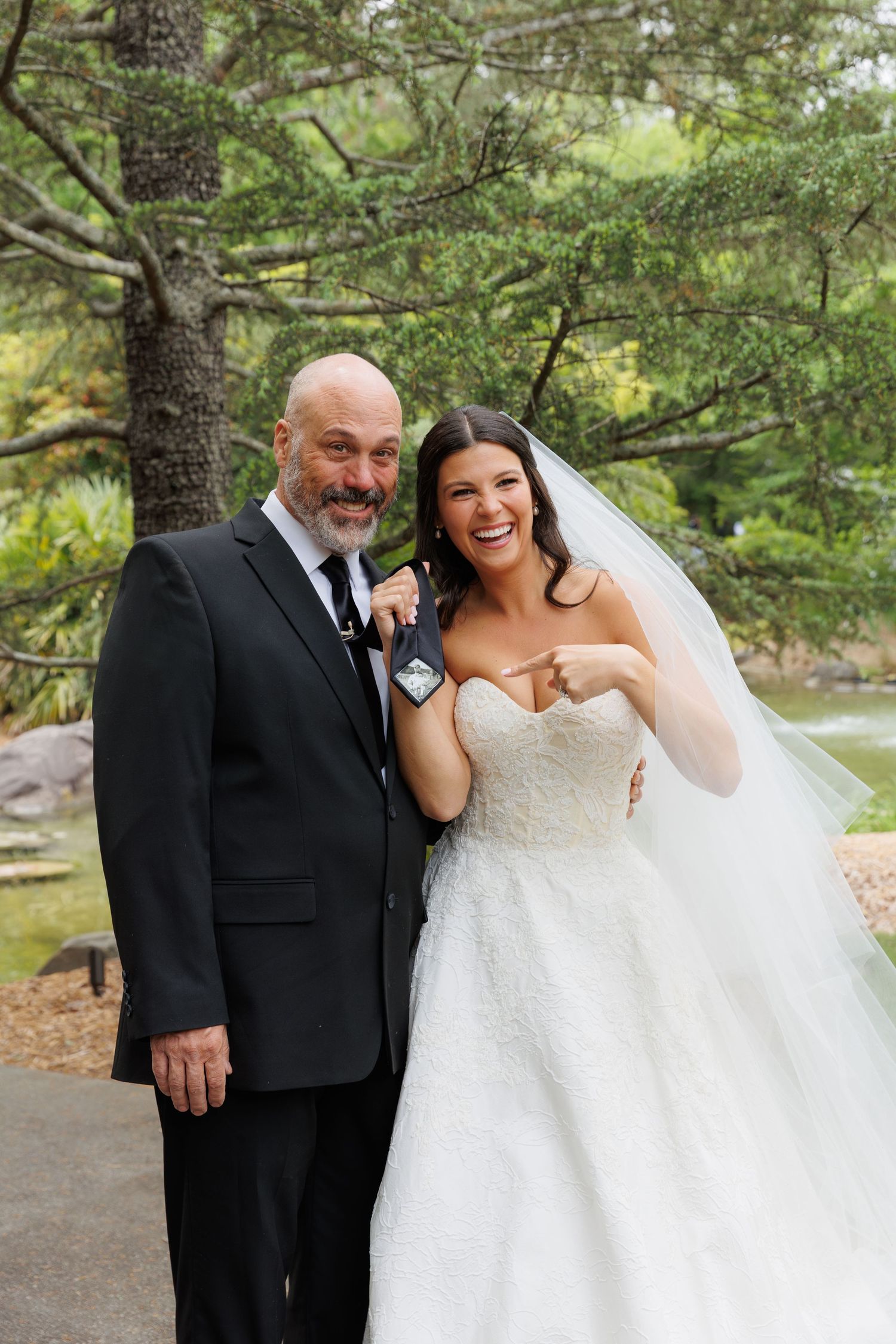 Bride in strapless ball gown laughs joyfully with older man in black tuxedo at outdoor garden wedding.