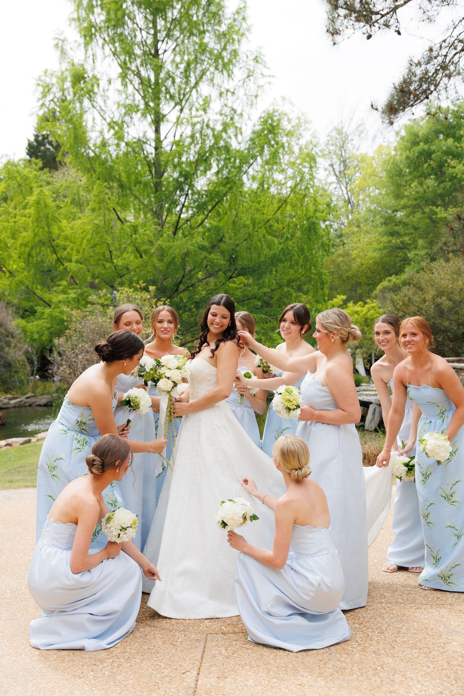 Bride in white gown surrounded by bridesmaids in light blue strapless dresses holding white bouquets in a garden setting.