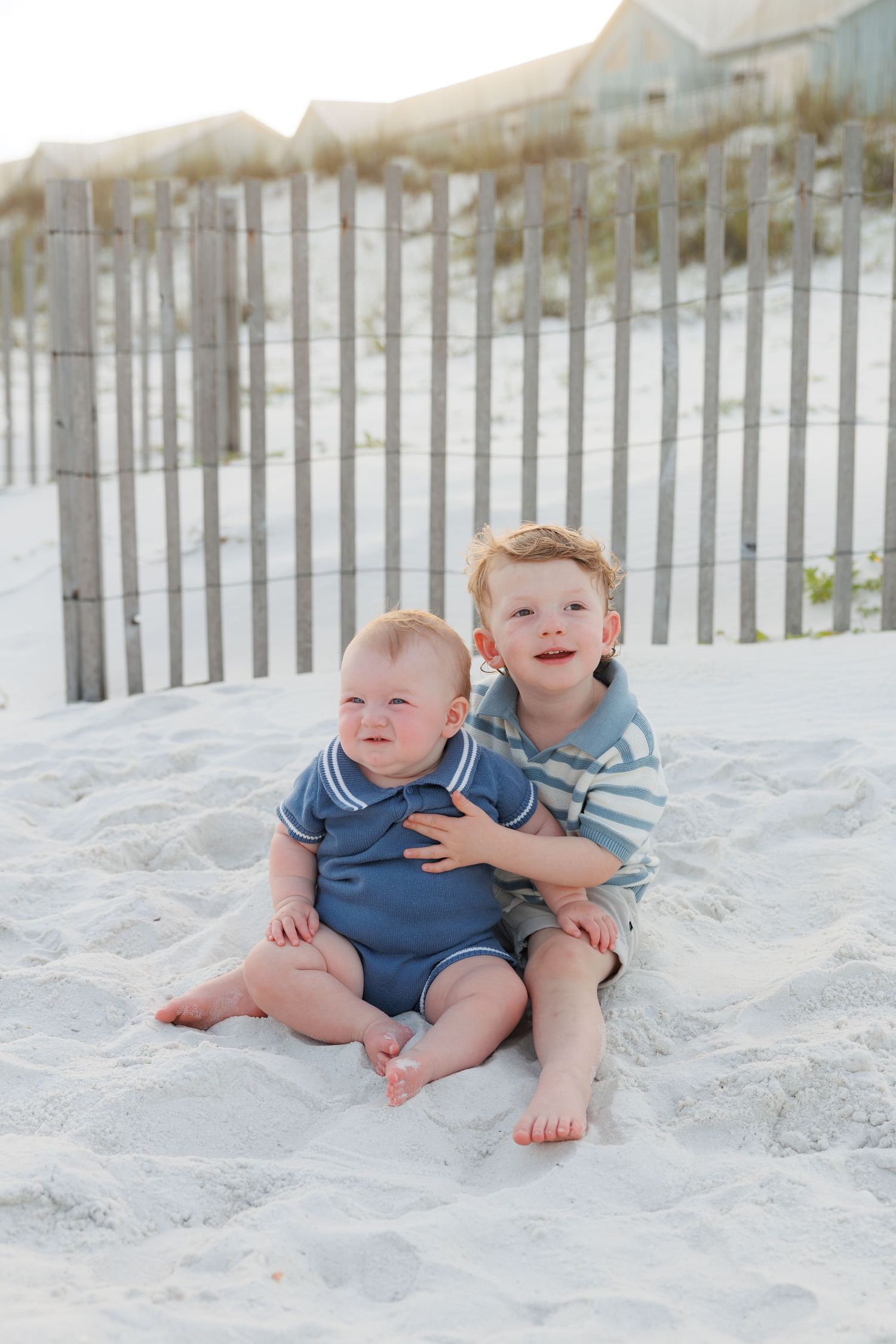 Two young brothers sitting together on white sand beach, older boy hugging infant sibling.