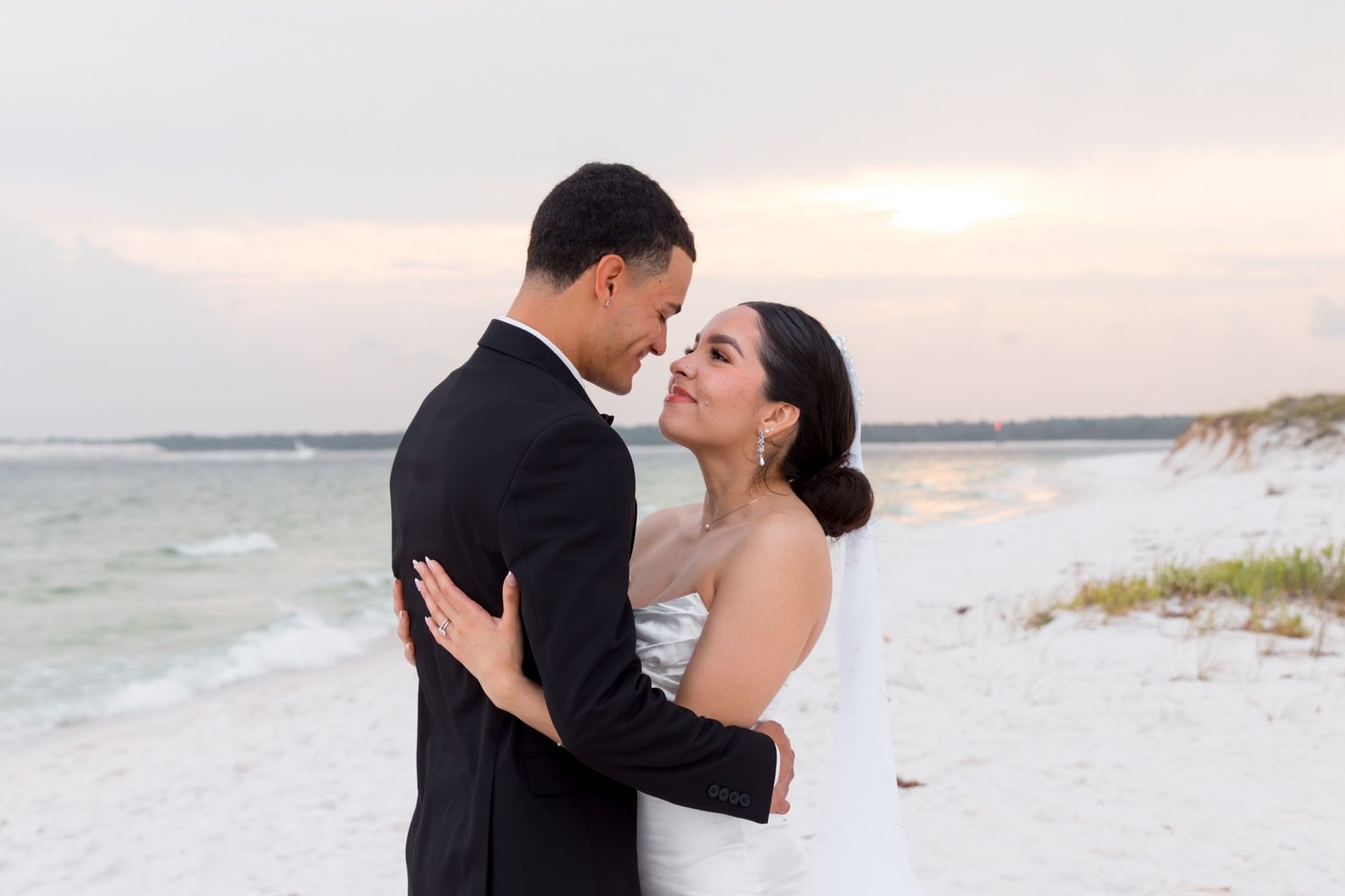 A couple in wedding attire share an intimate moment on a beach at sunset.
