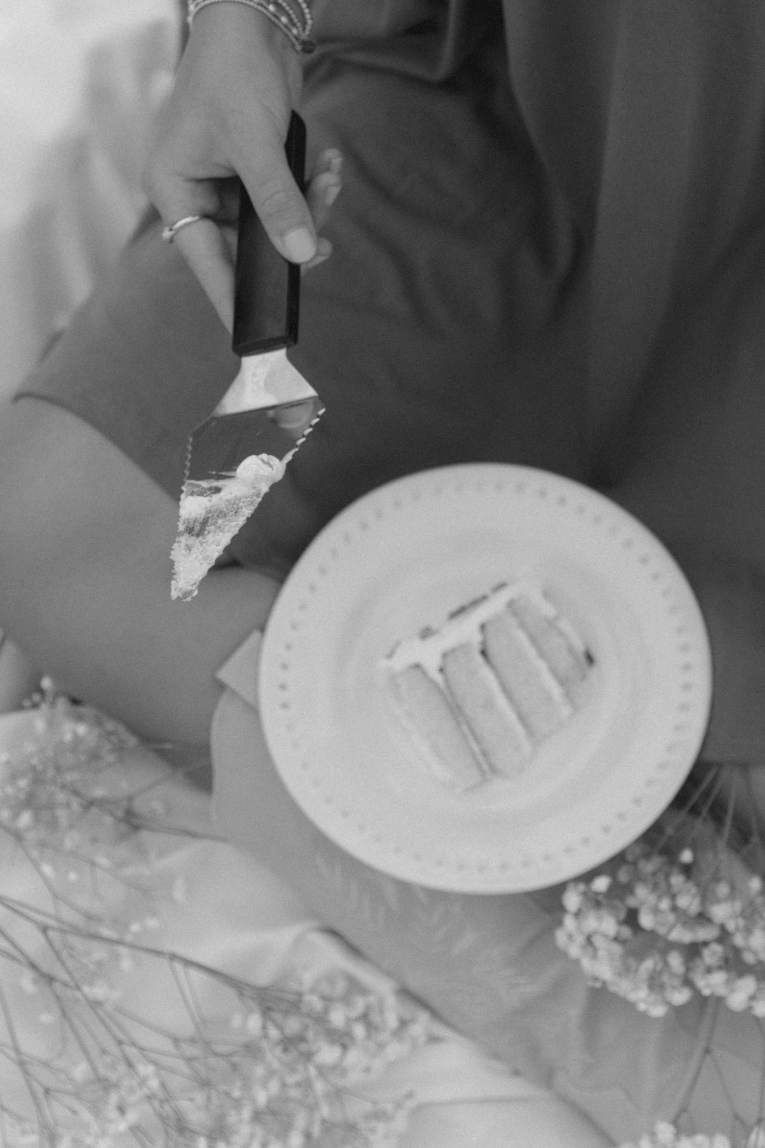 Black and white image of slicing a cake on a decorative plate with flowers nearby.
