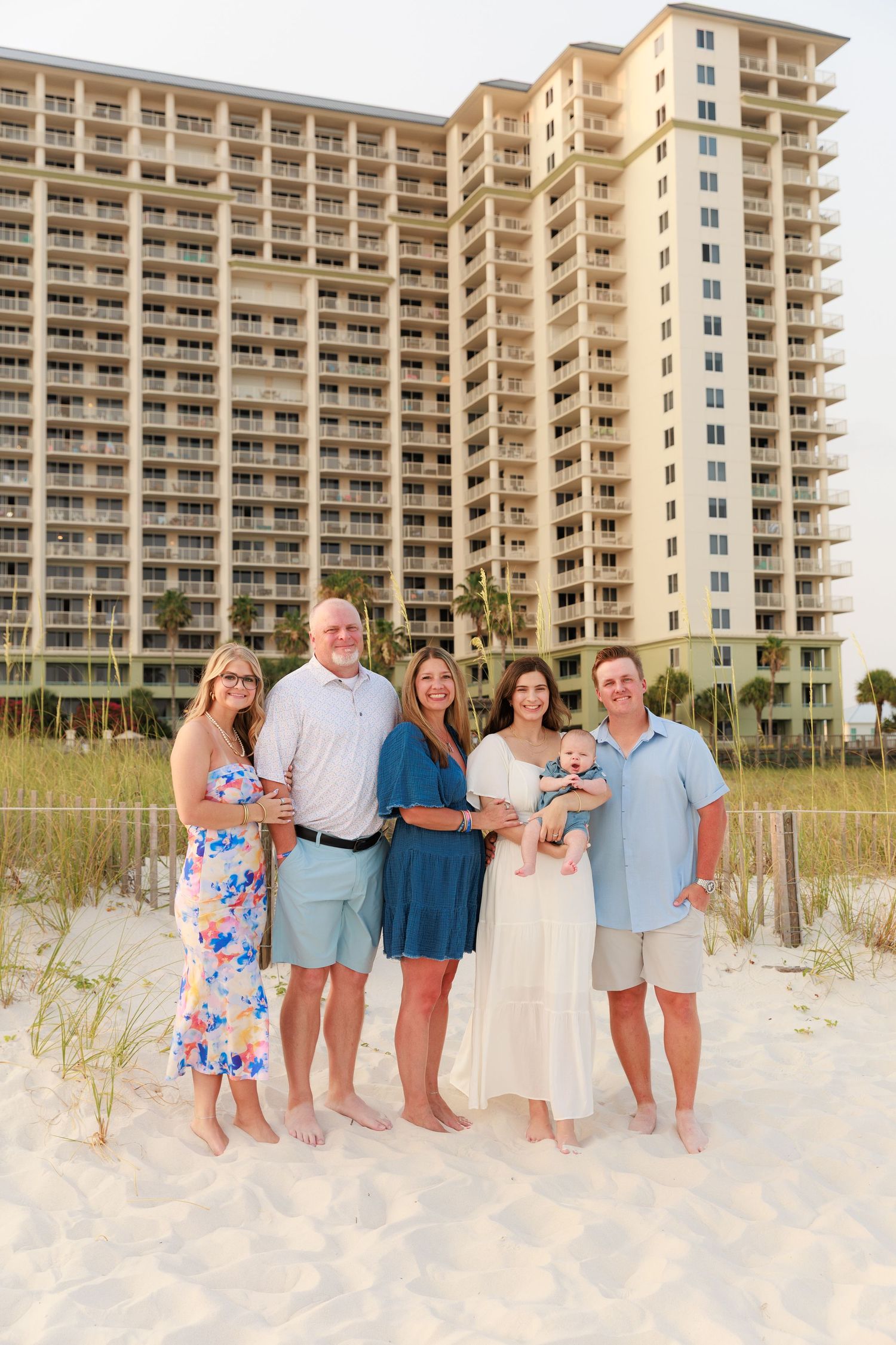 Family of six poses on sandy beach in front of large resort hotel during golden hour.