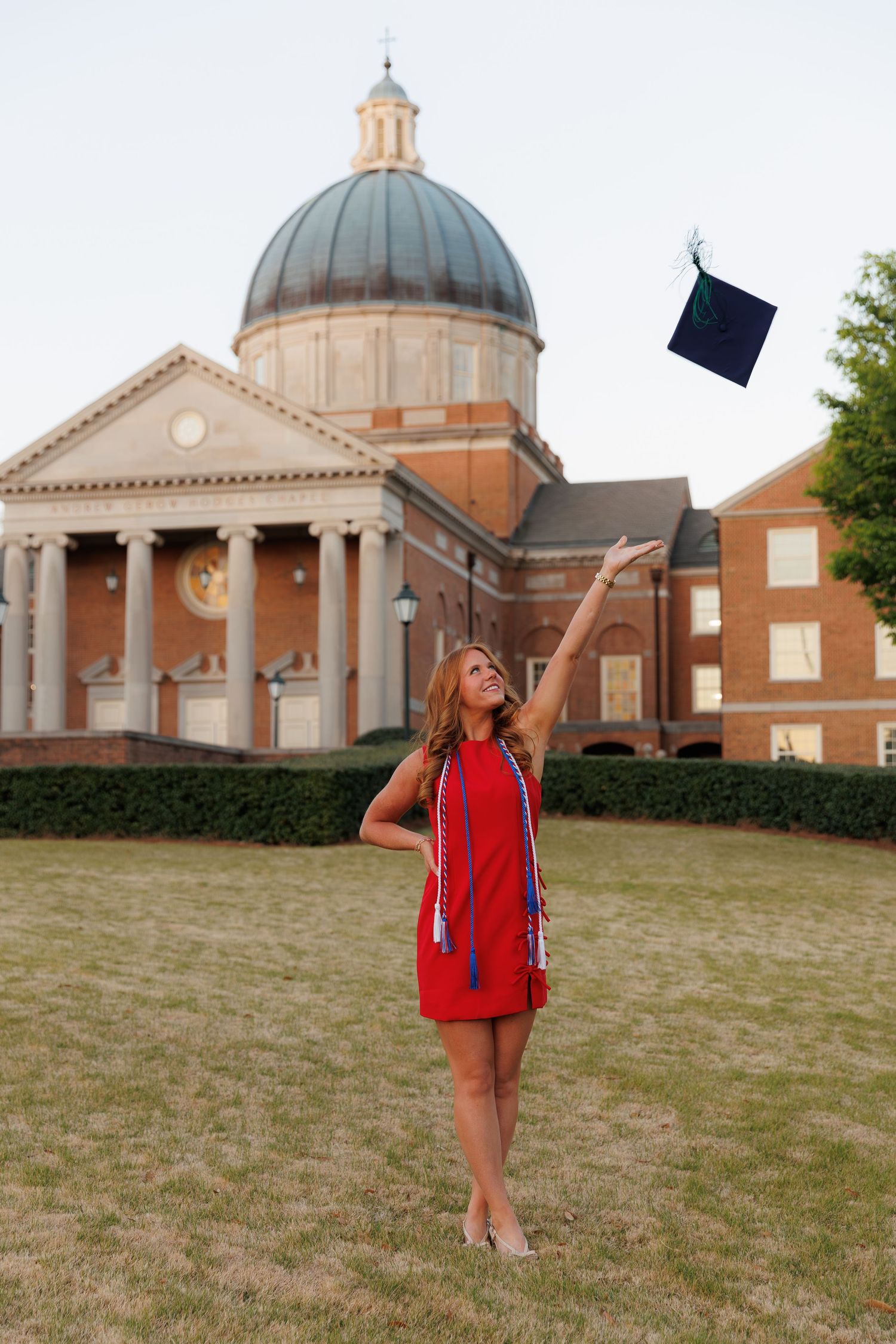 Young woman in red dress tossing graduation cap in front of a domed university building on a sunny day.