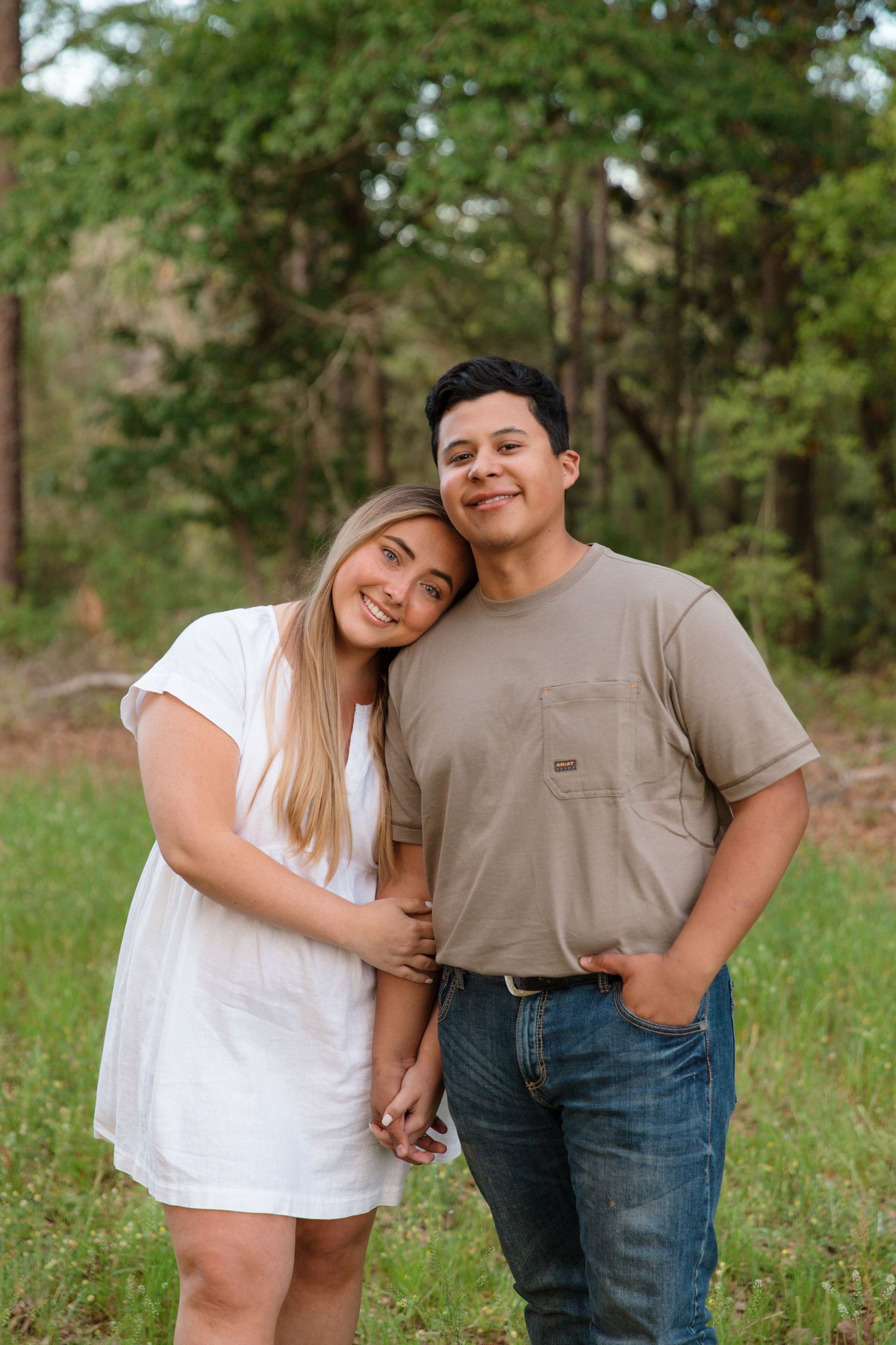 A young couple smiles together outdoors, woman in white dress leaning on man in tan shirt and jeans, green trees behind them.