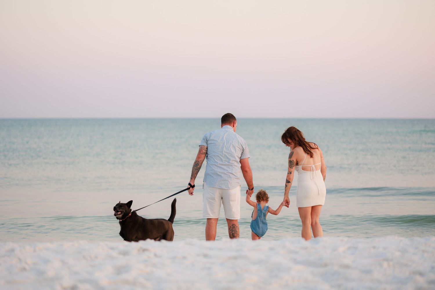 Family with dog walking on beach at sunset, parents holding child's hands near ocean waves.