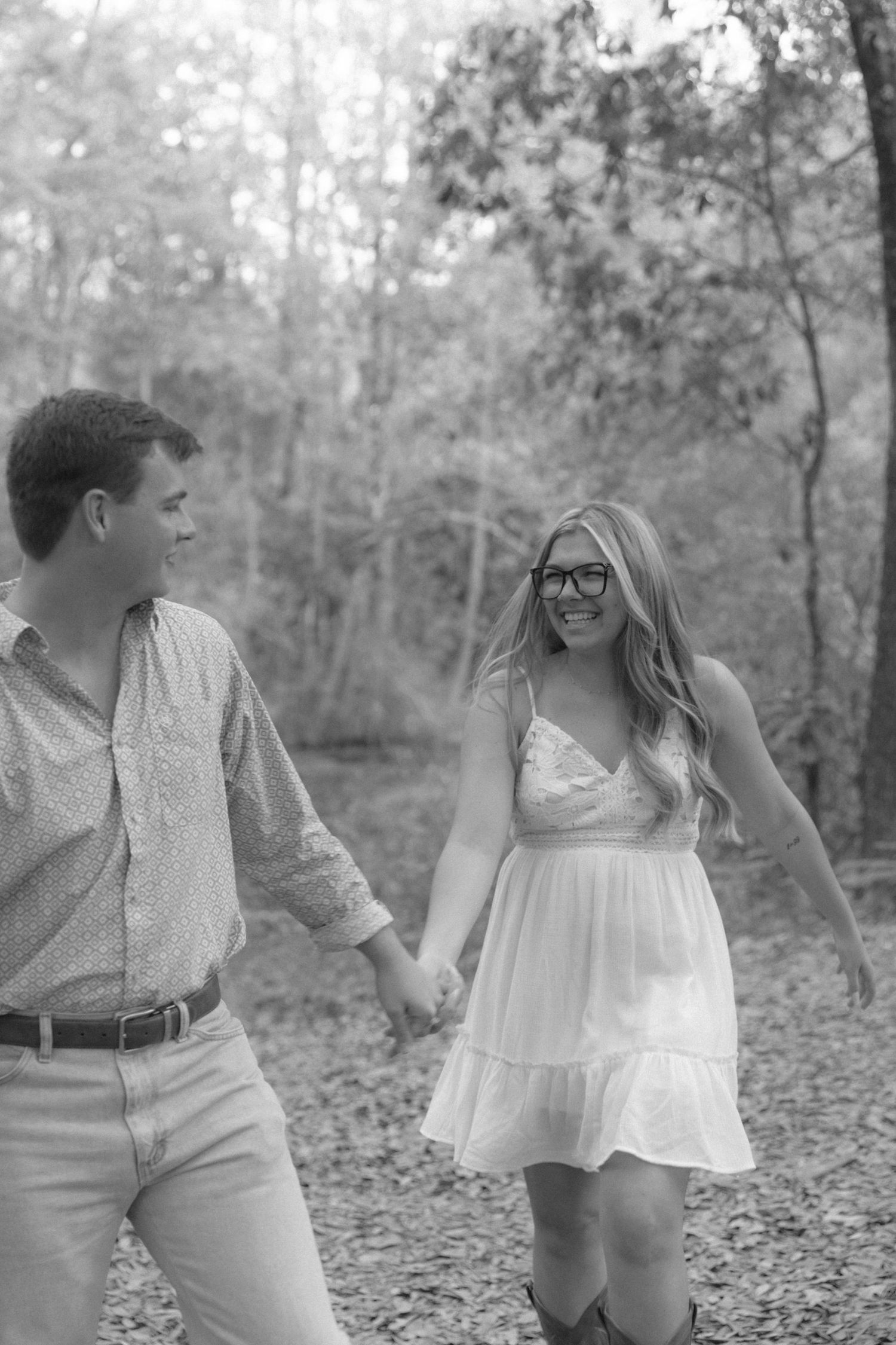 Black and white photo of a couple walking hand in hand through a wooded path, smiling at each other.