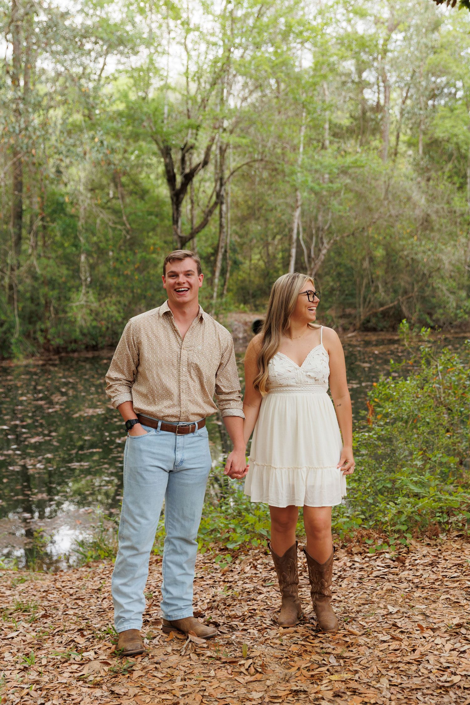 A smiling couple holds hands near a pond in a lush forest, dressed in casual country-style outfits.
