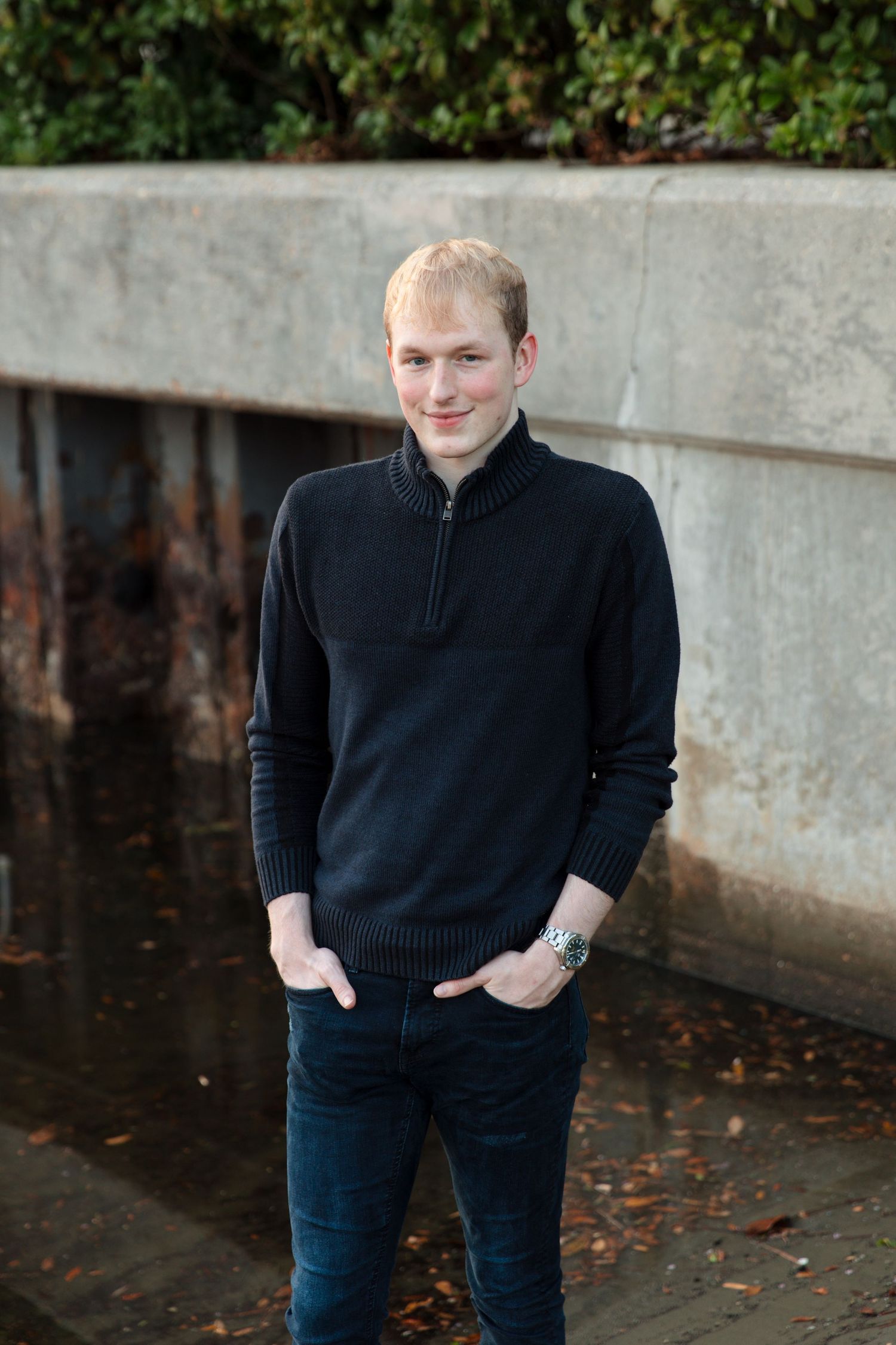 Young man in black zip-up sweater and jeans standing outdoors near a concrete wall with greenery.