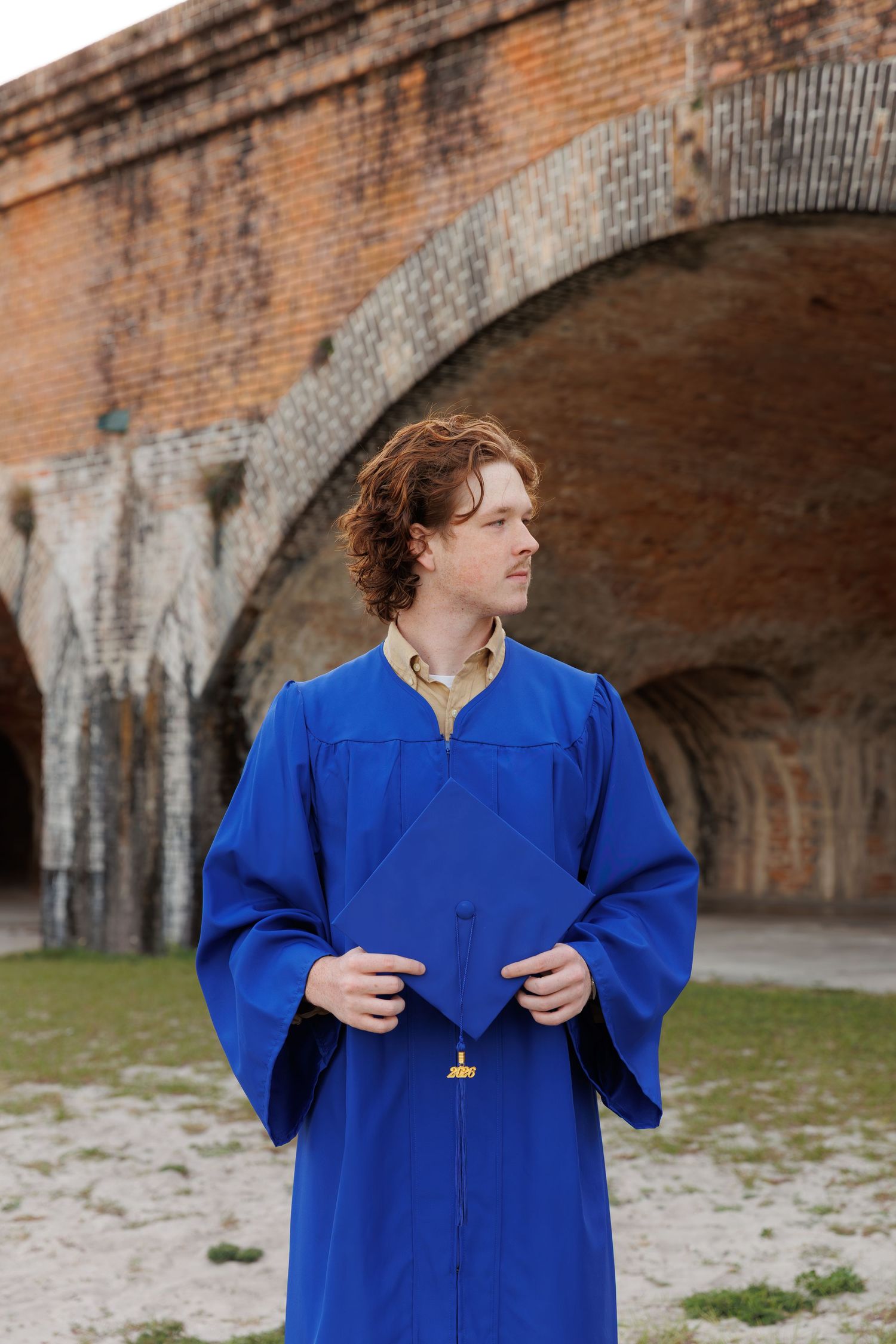 Young man in blue graduation gown holding cap before brick arches.