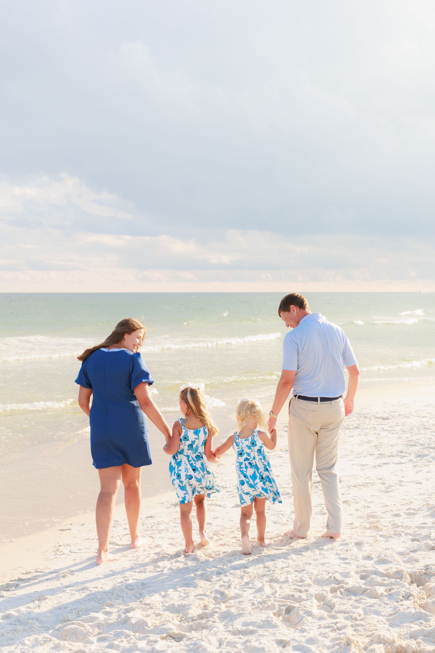 Family of four holding hands walking toward ocean waves on a white sandy beach, wearing coordinating blue outfits.