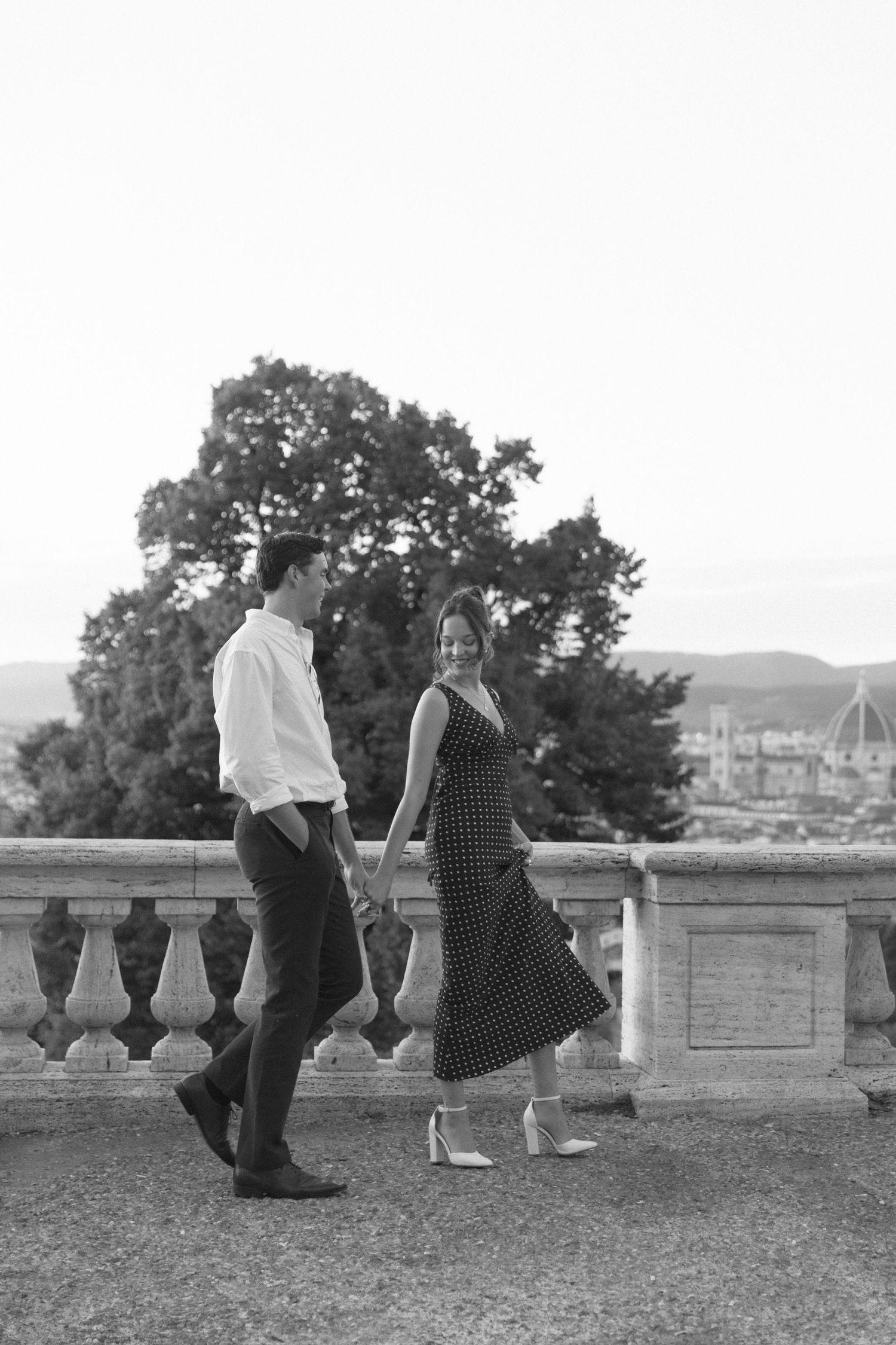 Black and white photo of a couple holding hands walking on a terrace with a scenic cityscape and trees in the background.