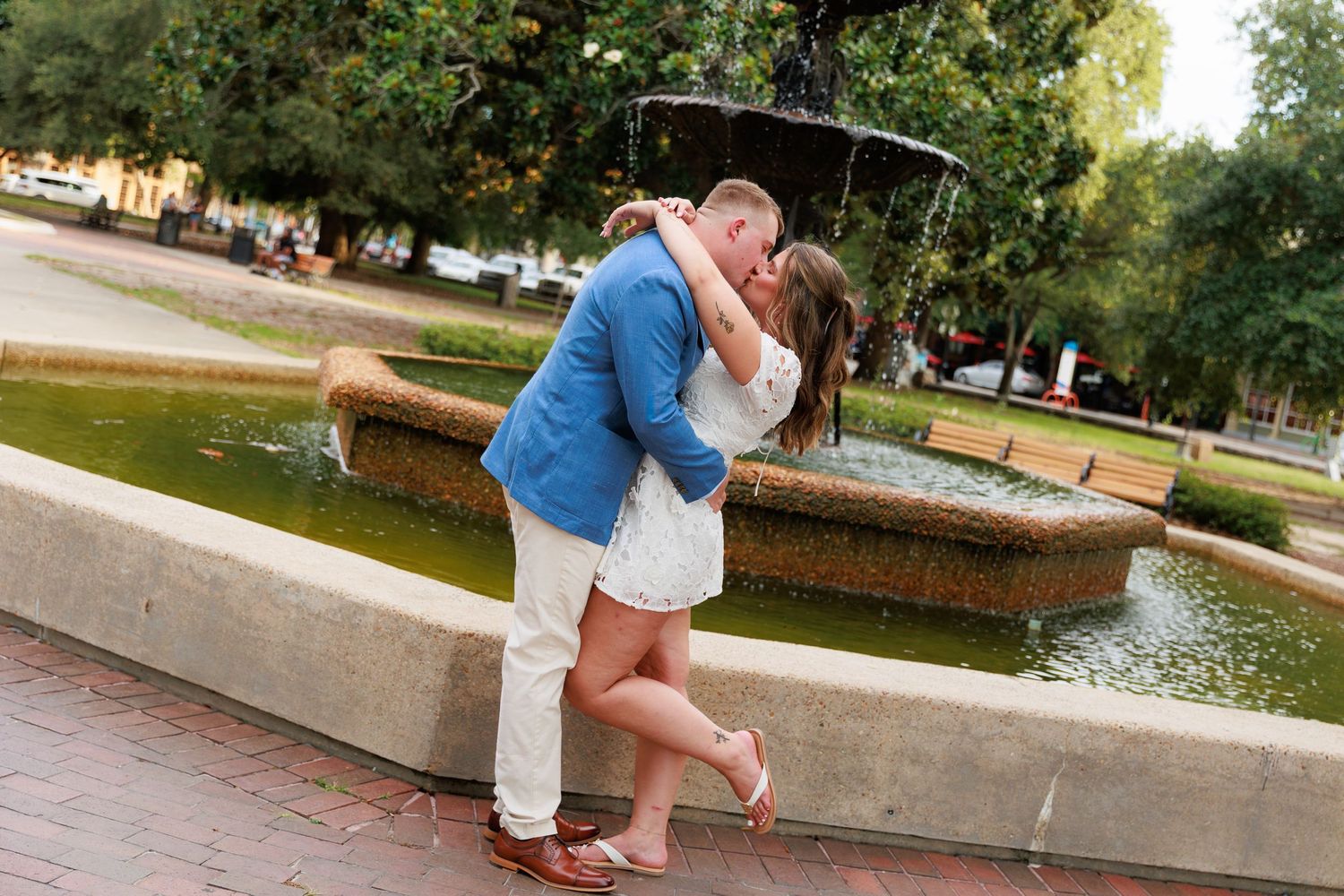 Couple sharing a romantic kiss in front of a decorative fountain in a park setting, man in blue blazer.