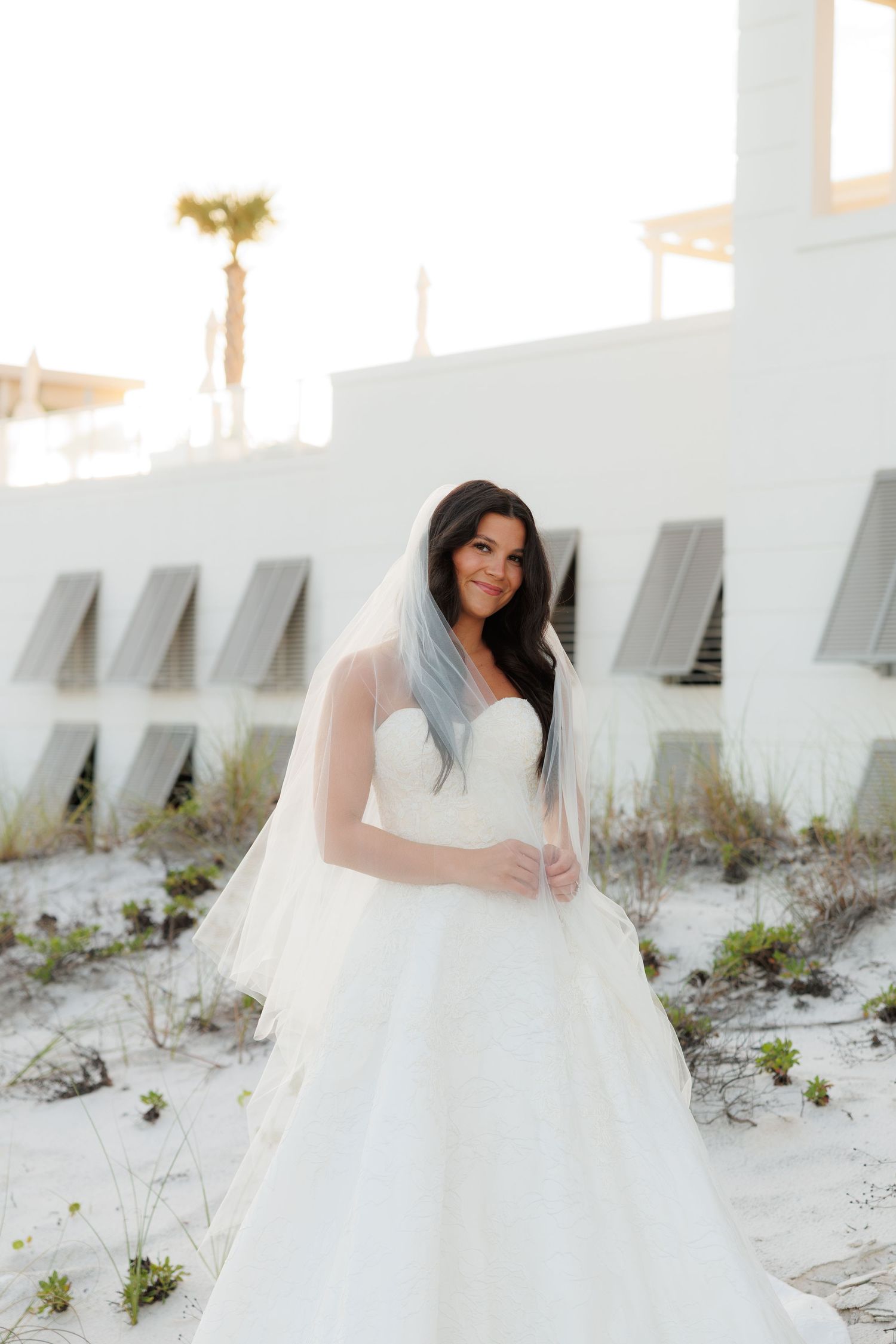 Bride in strapless white ball gown with veil poses on sandy beach near white building, long dark hair flowing.