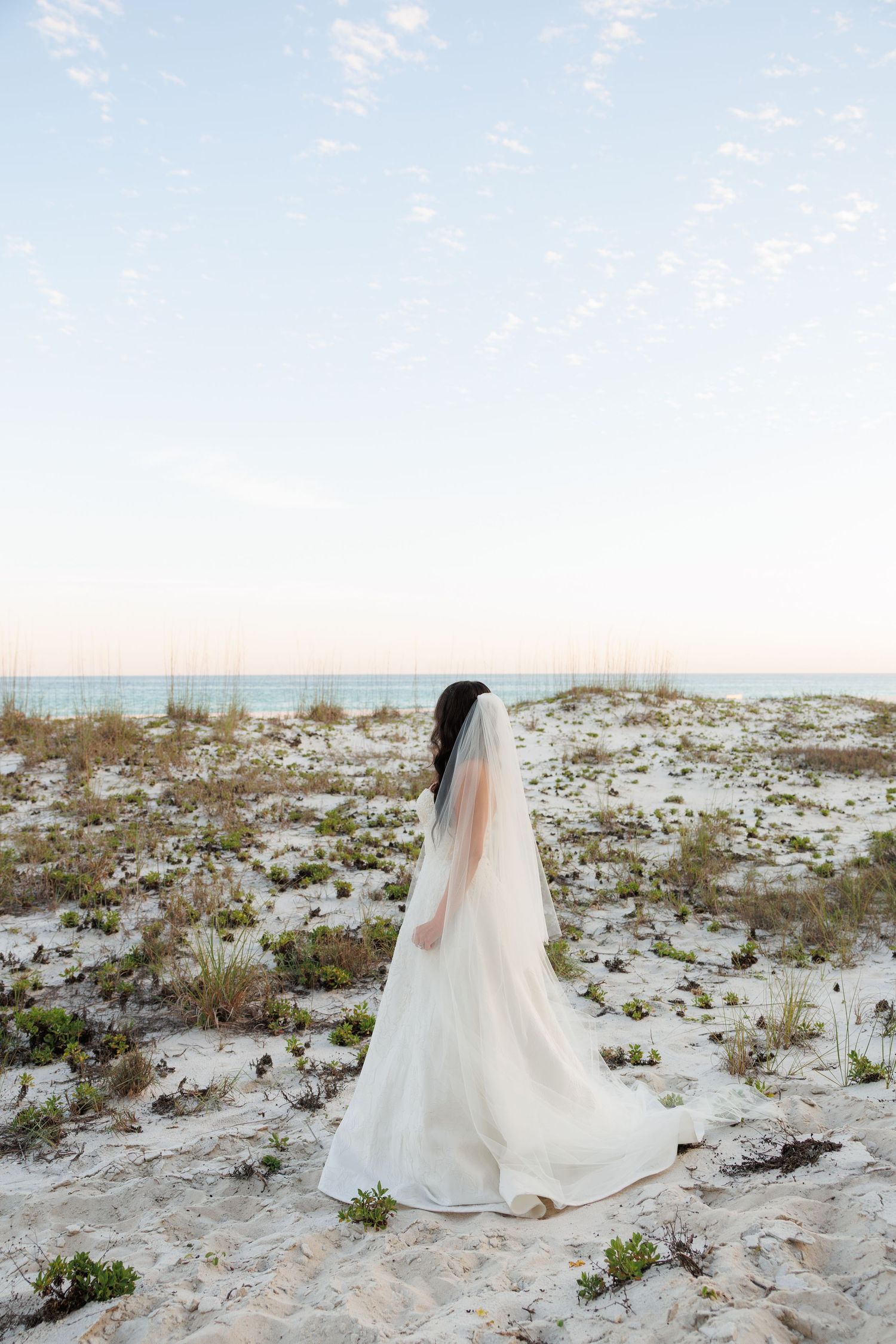 Bride in white wedding gown and long veil stands facing the ocean on a white sand beach at dusk.