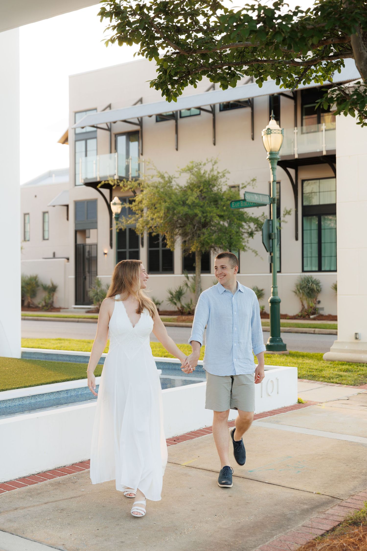 Couple holding hands walking along a sunny sidewalk, woman in white dress, man in light blue shirt and gray shorts.
