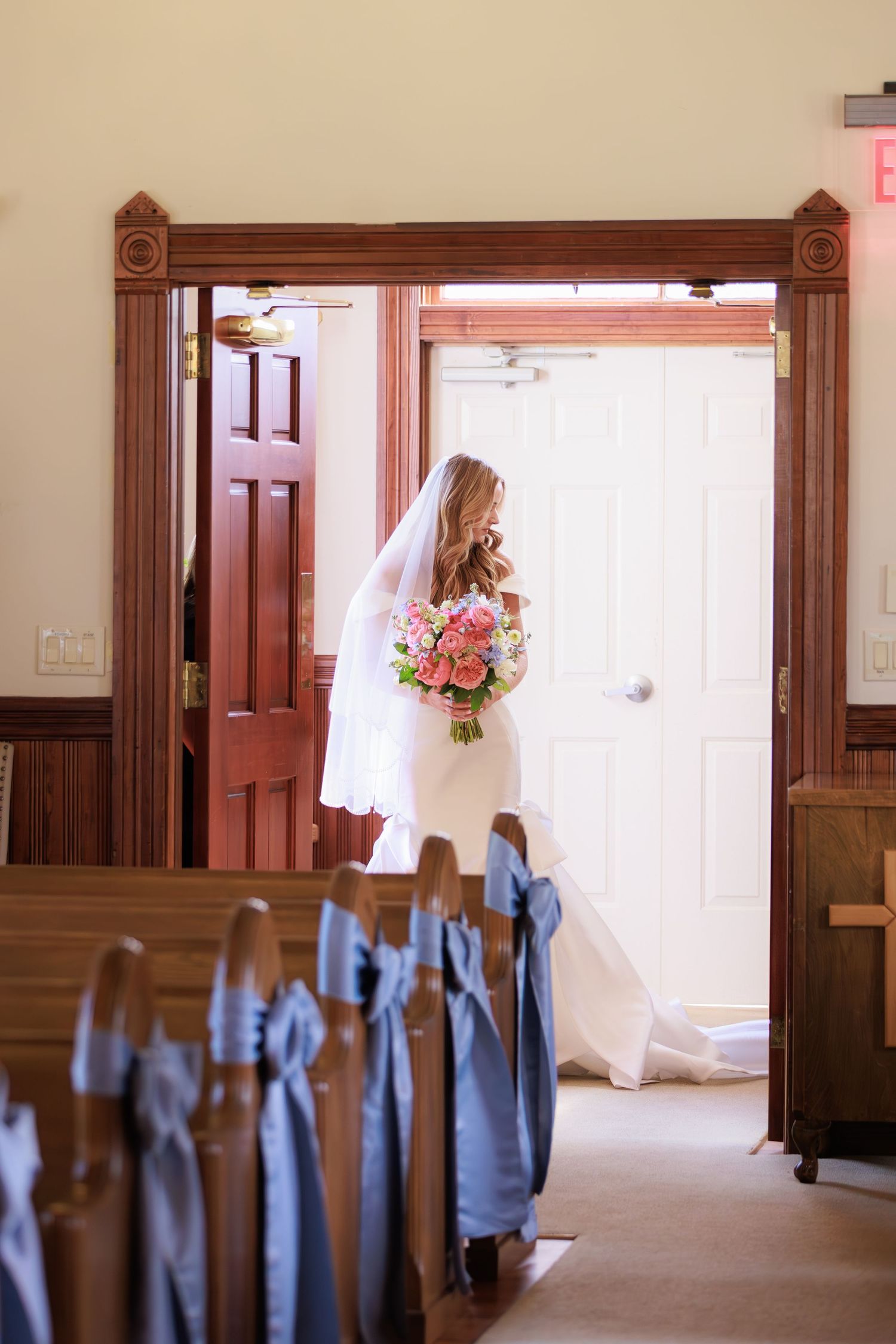 A bride in white dress holding a pink bouquet stands in a wooden church doorway framed by traditional pews.