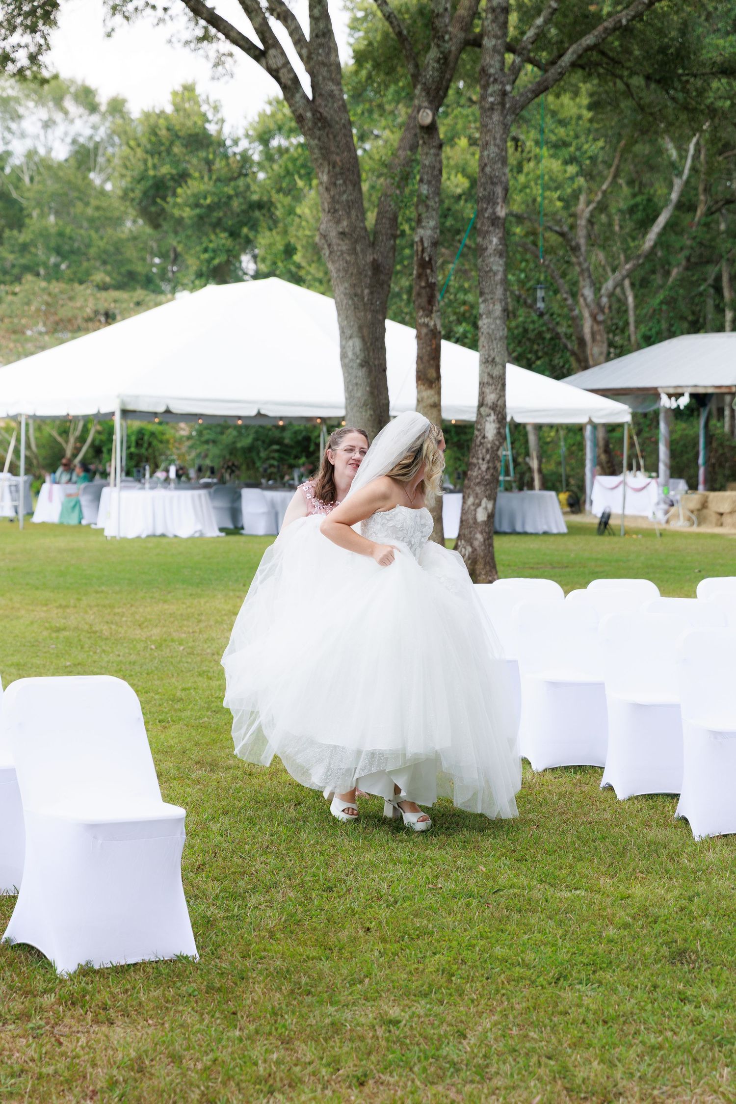 White-covered chairs arranged on a grassy lawn for an elegant outdoor wedding reception under shady trees.