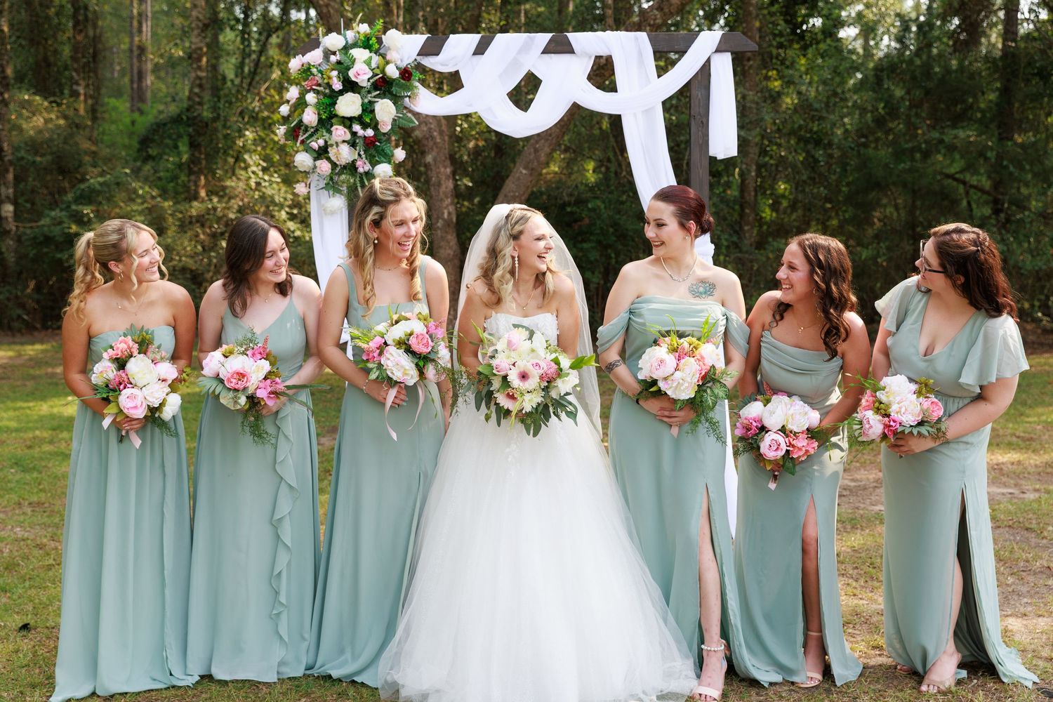 Wedding party wearing sage green bridesmaid dresses poses together with bouquets in an outdoor garden setting.
