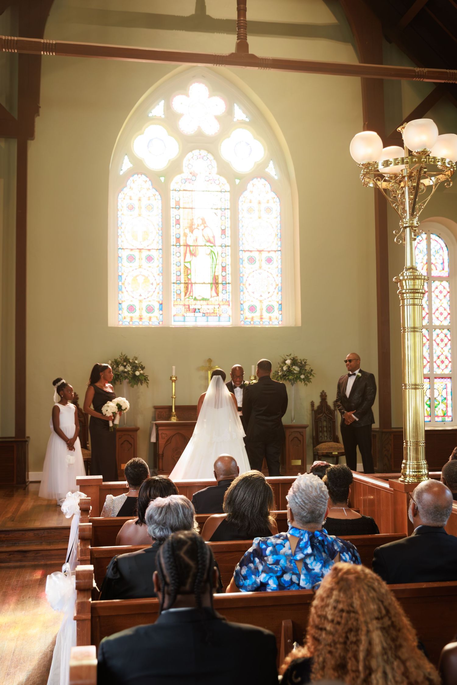 A church wedding ceremony in progress with stained glass windows and wooden pews filled with guests watching the proceedings.