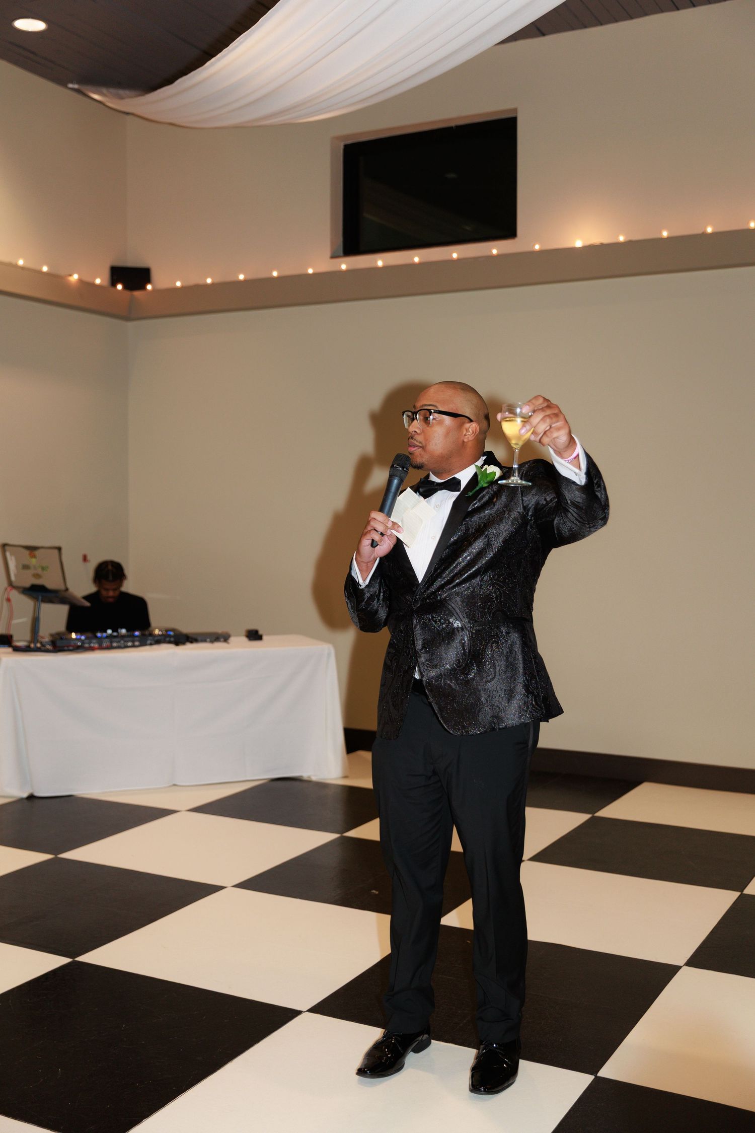 A person in a tuxedo gives a speech while holding a microphone on a checkered dance floor.
