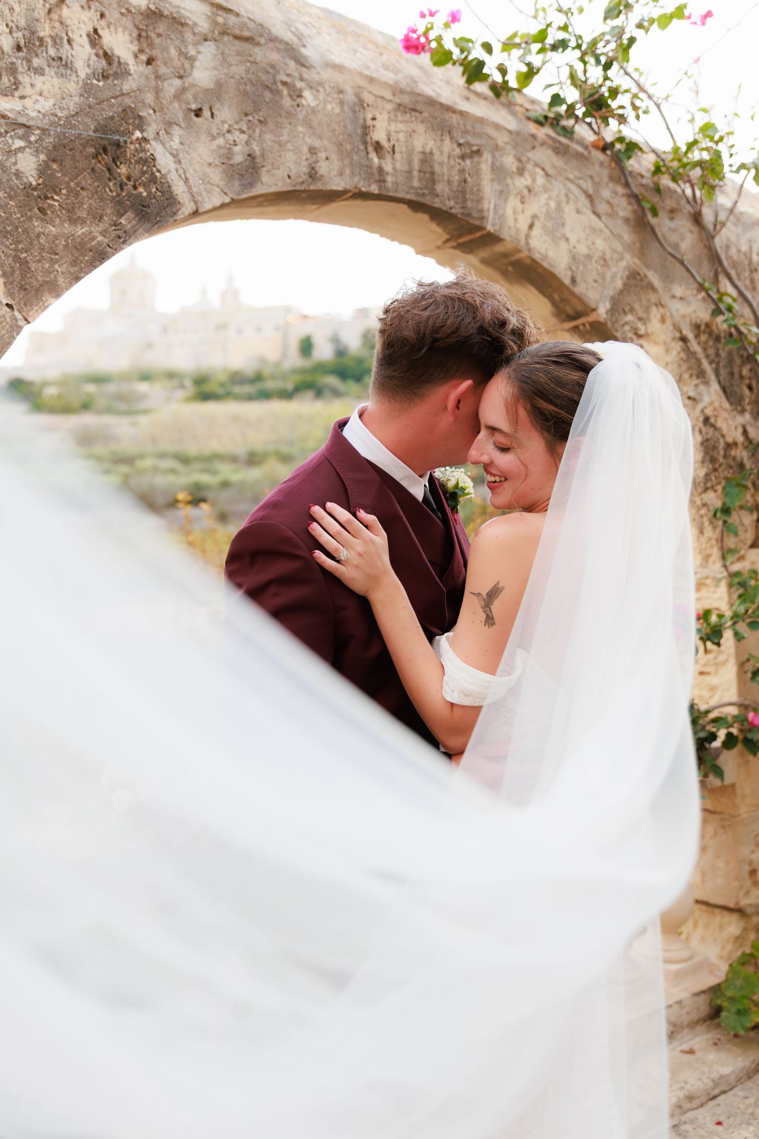 A tender wedding moment framed by a stone arch with flowing white veil in motion.