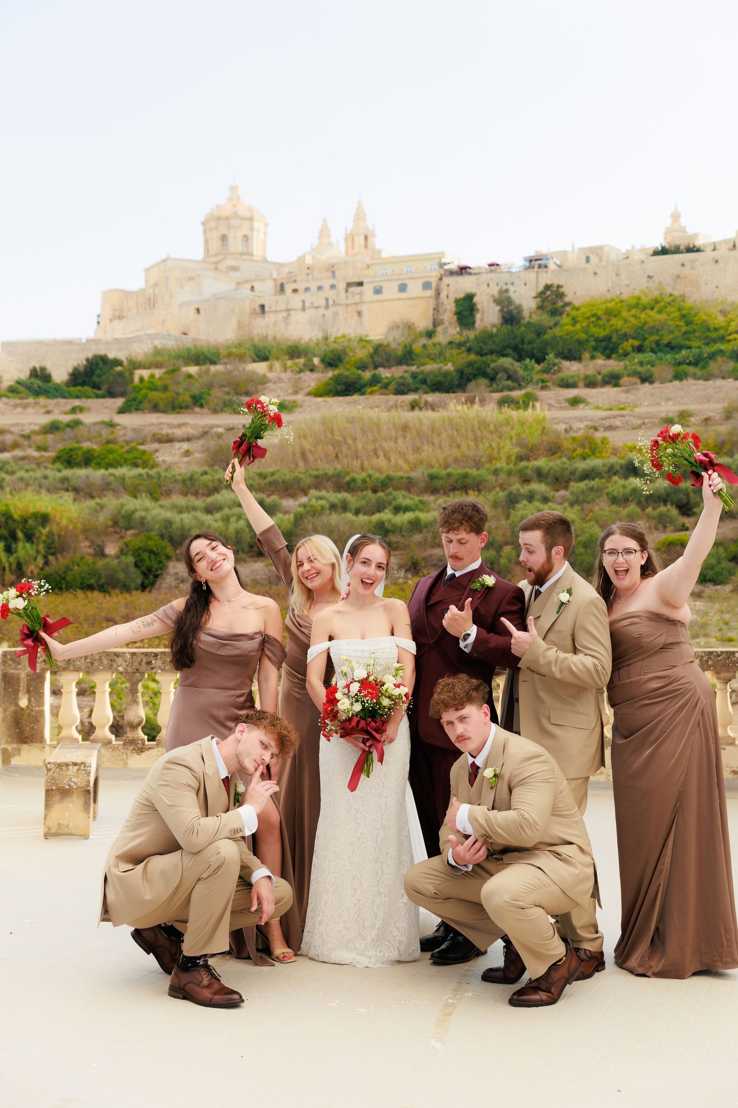 Wedding party poses together on terrace with historic Mediterranean cathedral and fortress buildings in background.