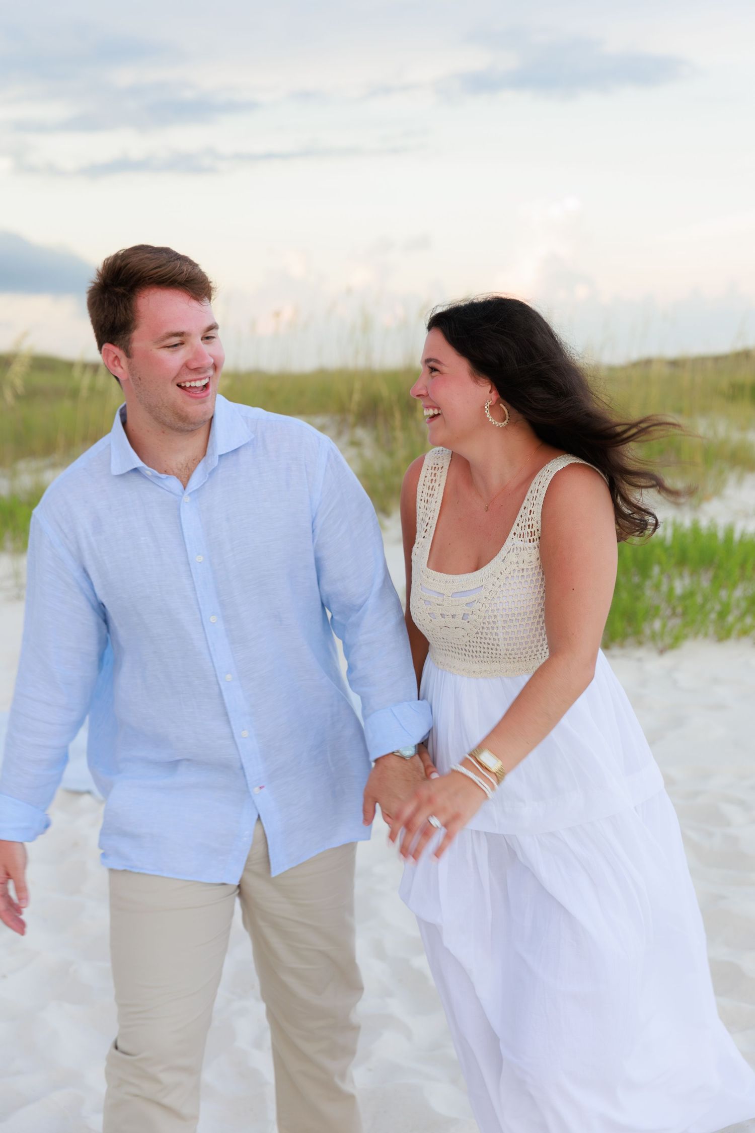 A couple laughs and walks together on a beach wearing coordinating light blue and white outfits.