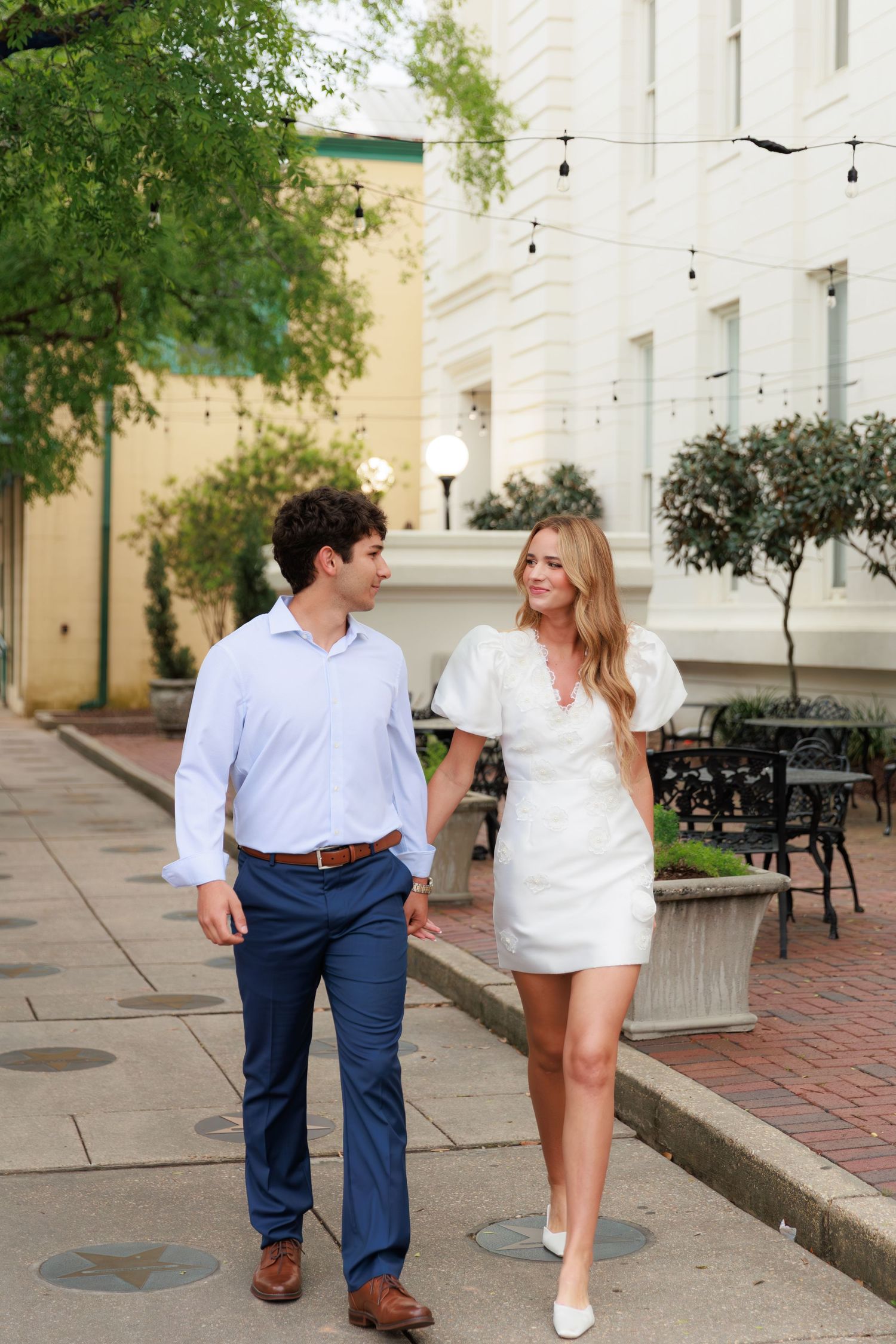 A couple walking and interacting playfully along a brick-paved walkway with string lights overhead.