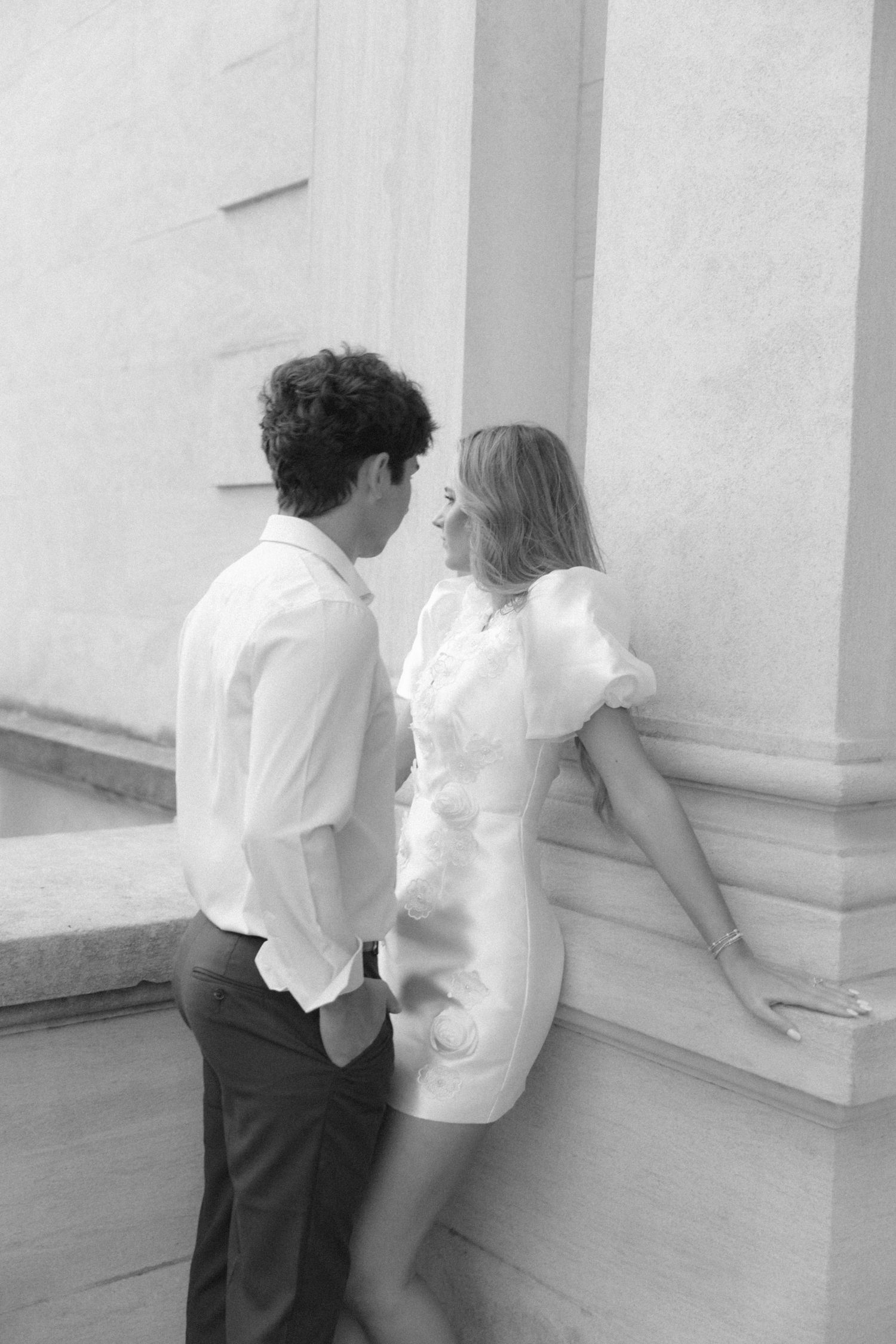 A black and white photo capturing a romantic moment between a couple against a white building exterior.