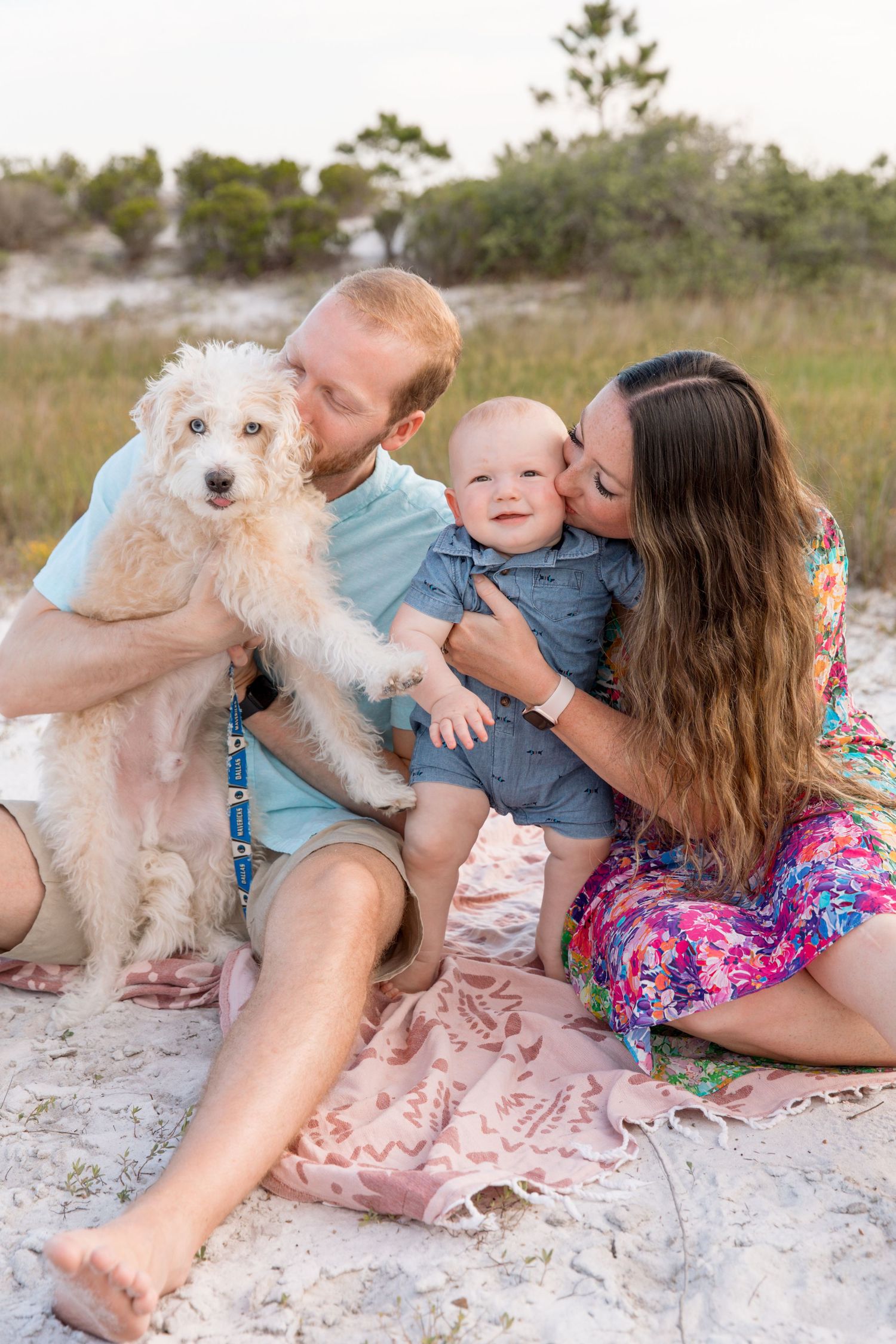 A family and their fluffy white dog share tender moments together on a sandy beach at sunset.