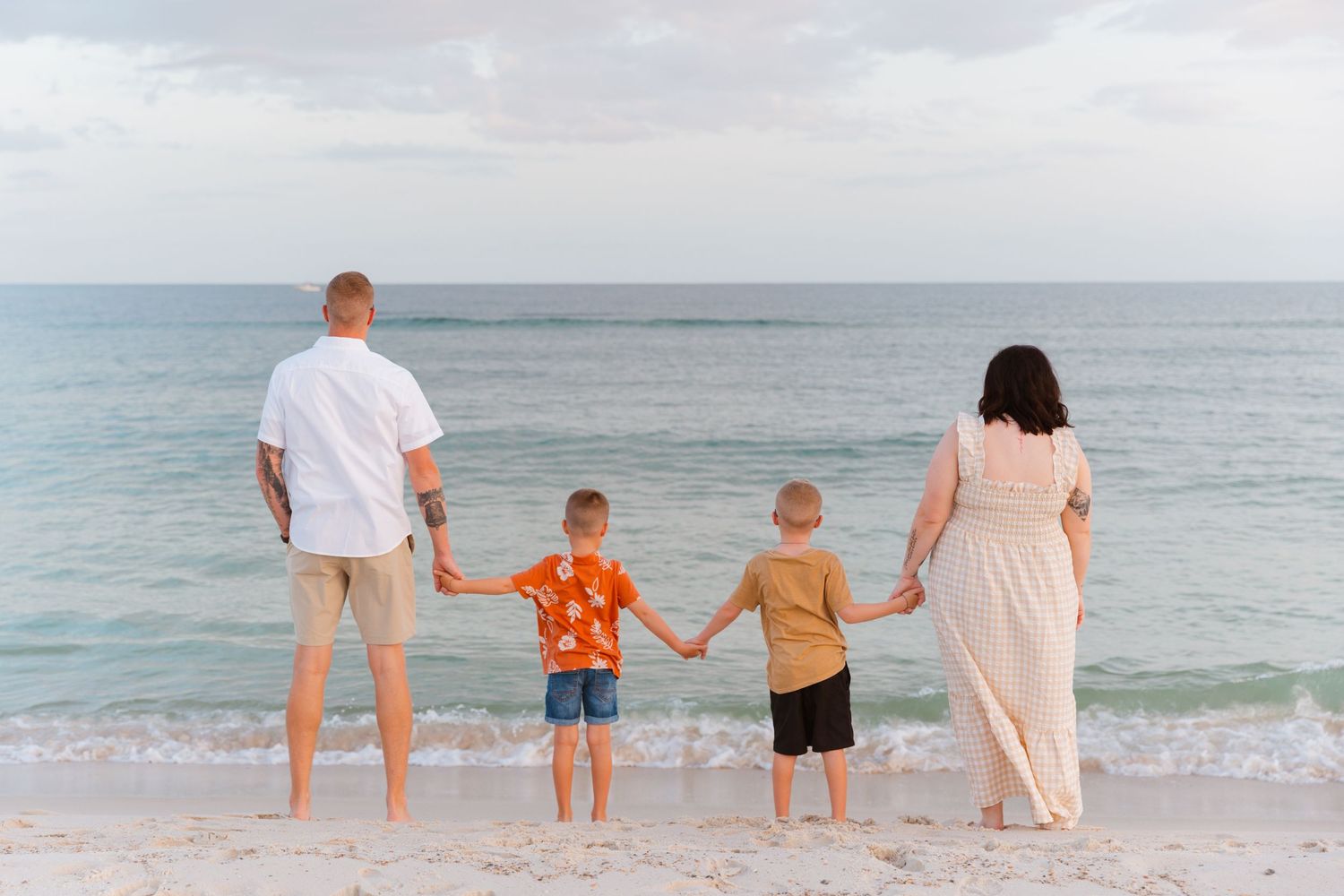 Family of four holds hands while looking at ocean from beach at sunset.